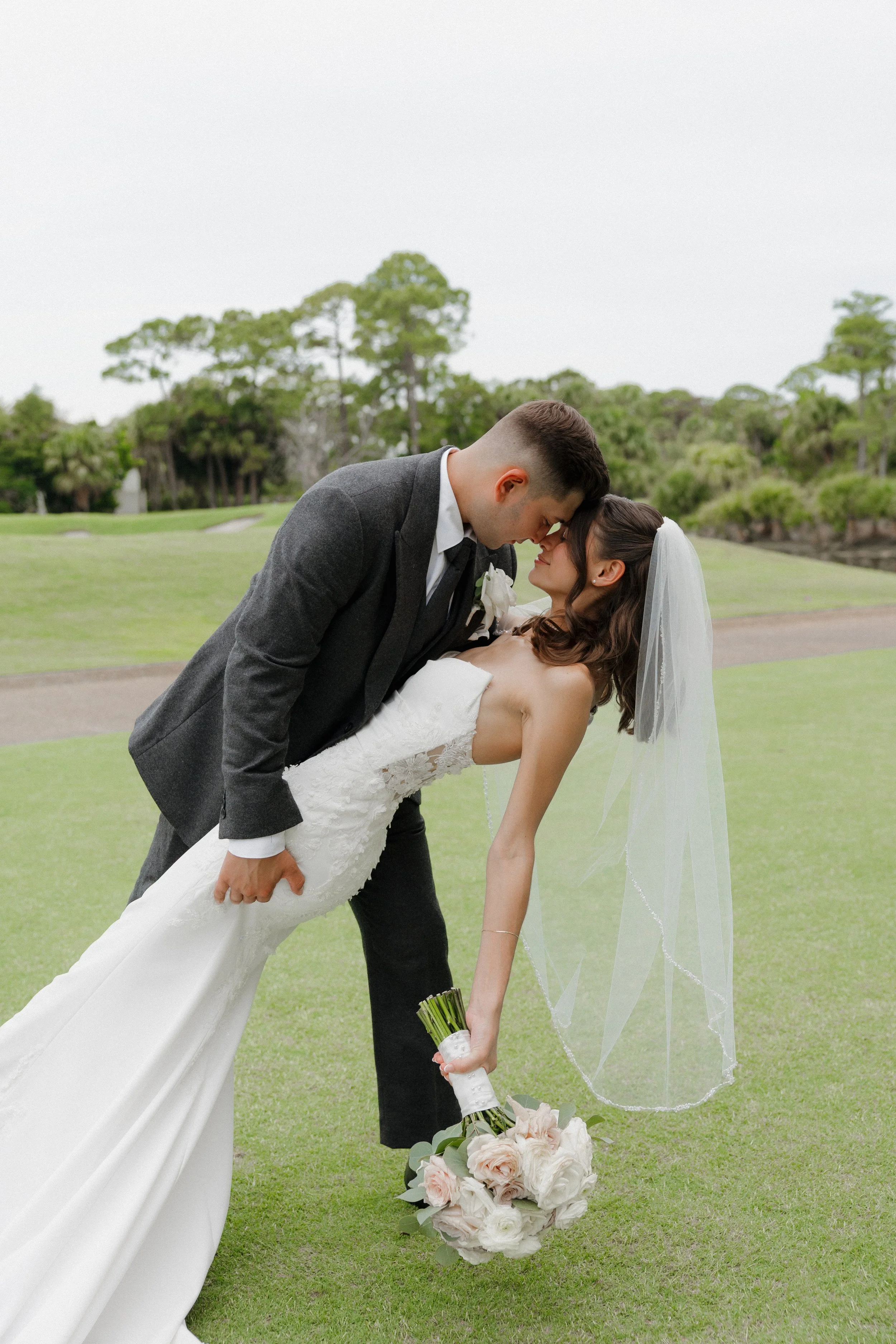 A bride and groom dancing outside on a grassy field during their wedding, with the groom dipping the bride while holding her waist and the bride holding a bouquet on the ground with a large floral arrangement.