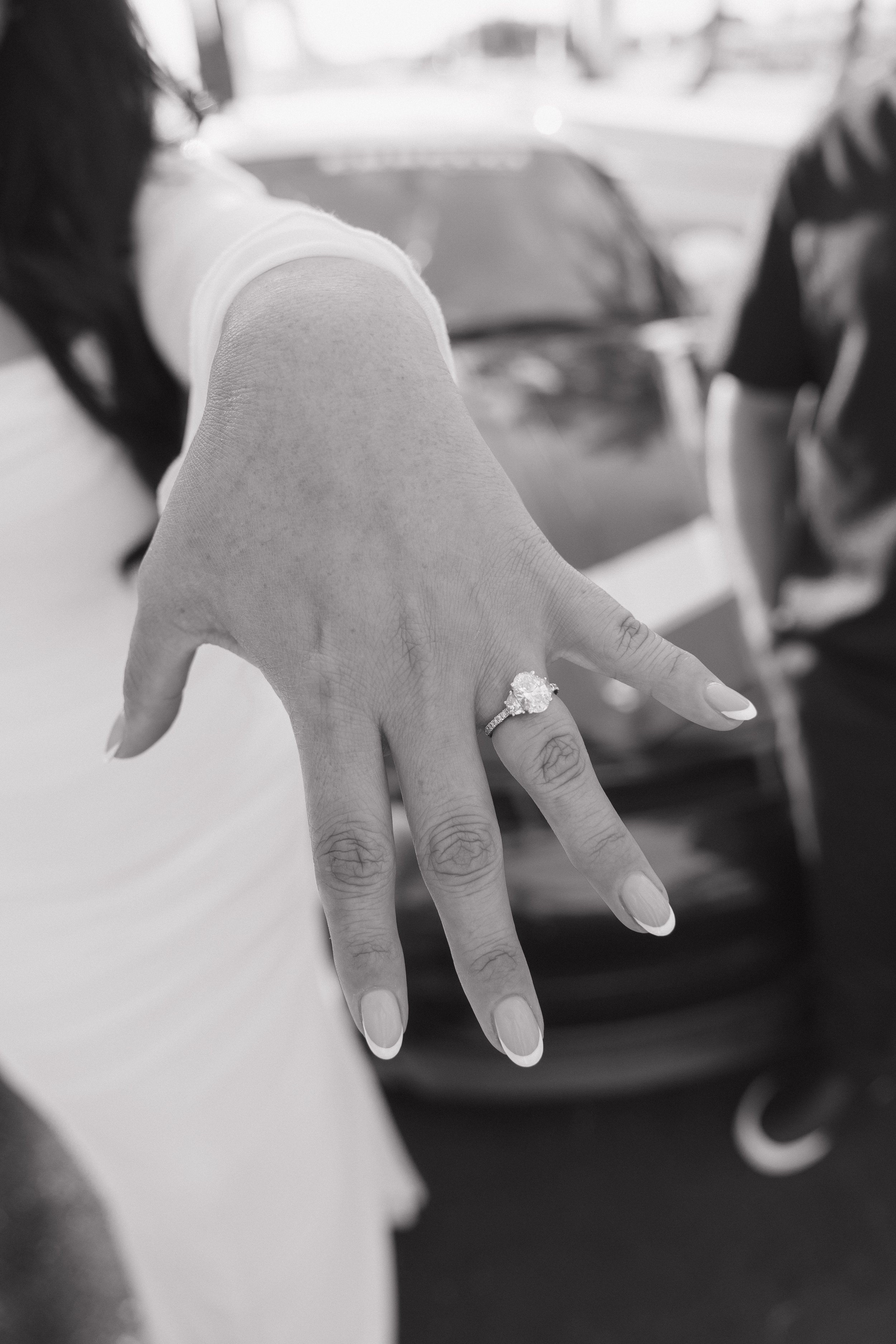 Close-up of a woman's hand showing an engagement ring with a large central diamond, with the background blurred and showing part of her hair and clothing.