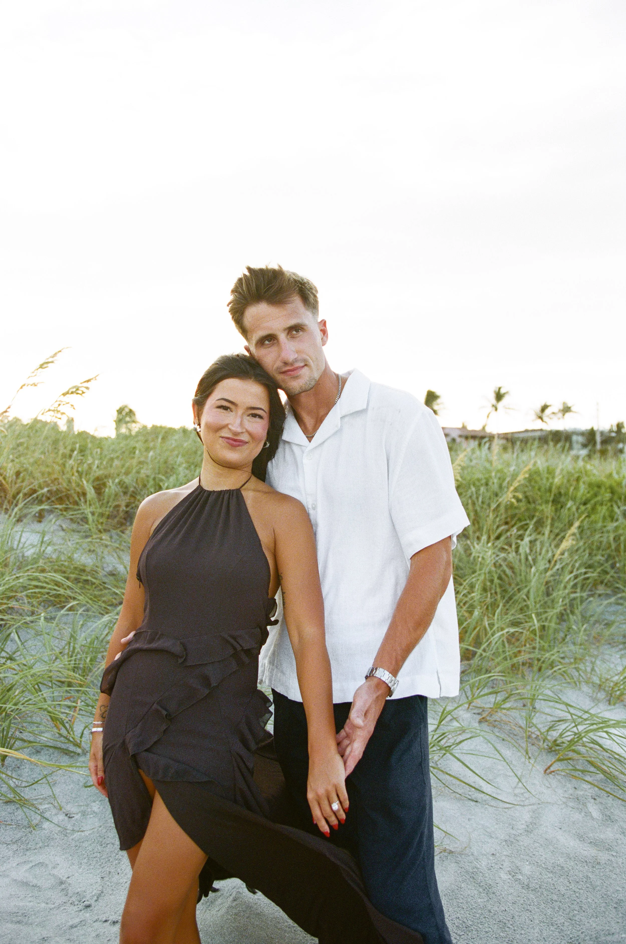 A couple standing together outdoors on a sandy path surrounded by green plants, with palm trees in the background, during daylight.