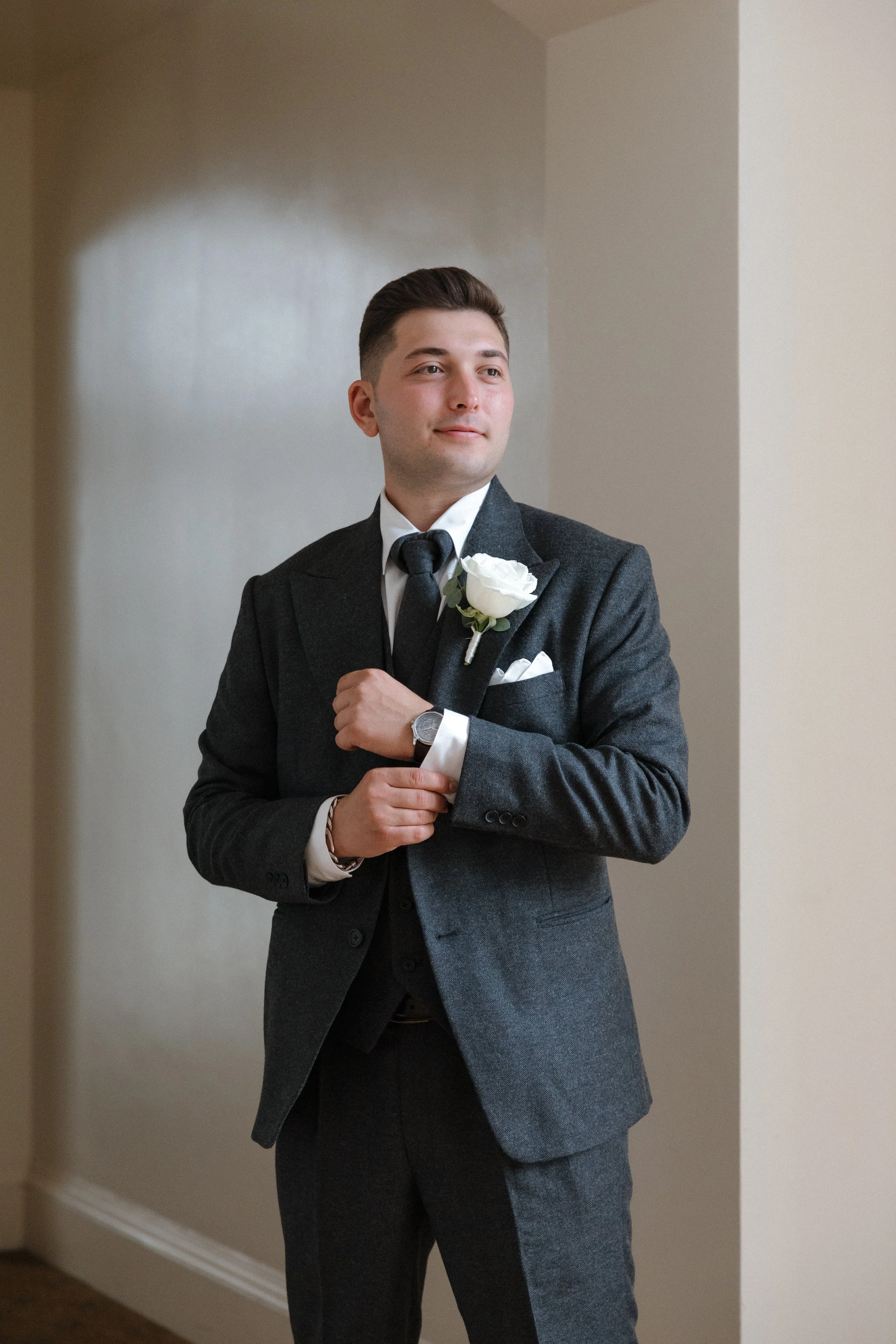 A young man in a dark suit with a white shirt and black tie, wearing a wristwatch and a boutonniere on his lapel, standing indoors and adjusting his cufflink, with neutral-colored walls in the background.