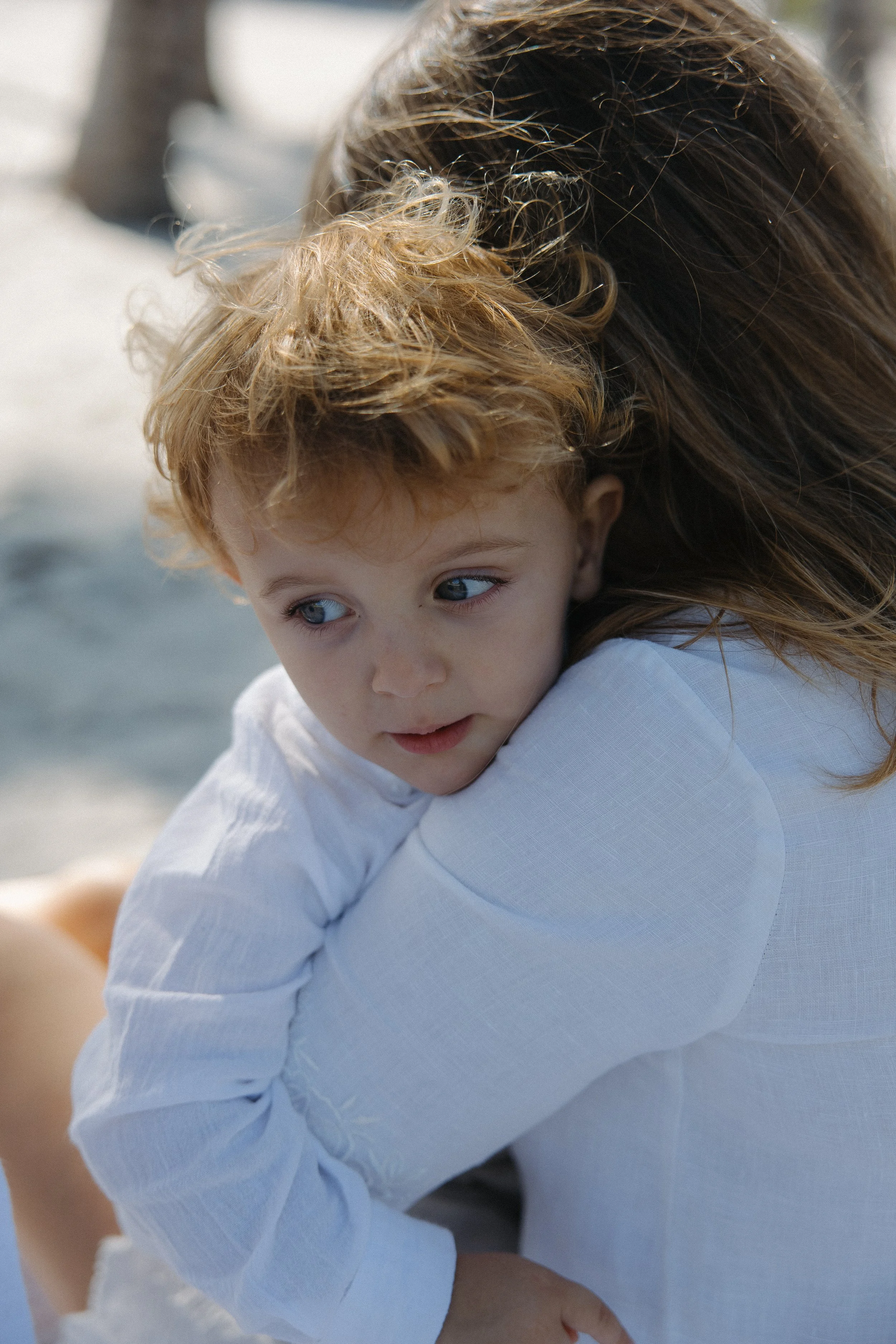 A close-up of a young child with blue eyes and curly, light brown hair, being held by an adult on a beach.