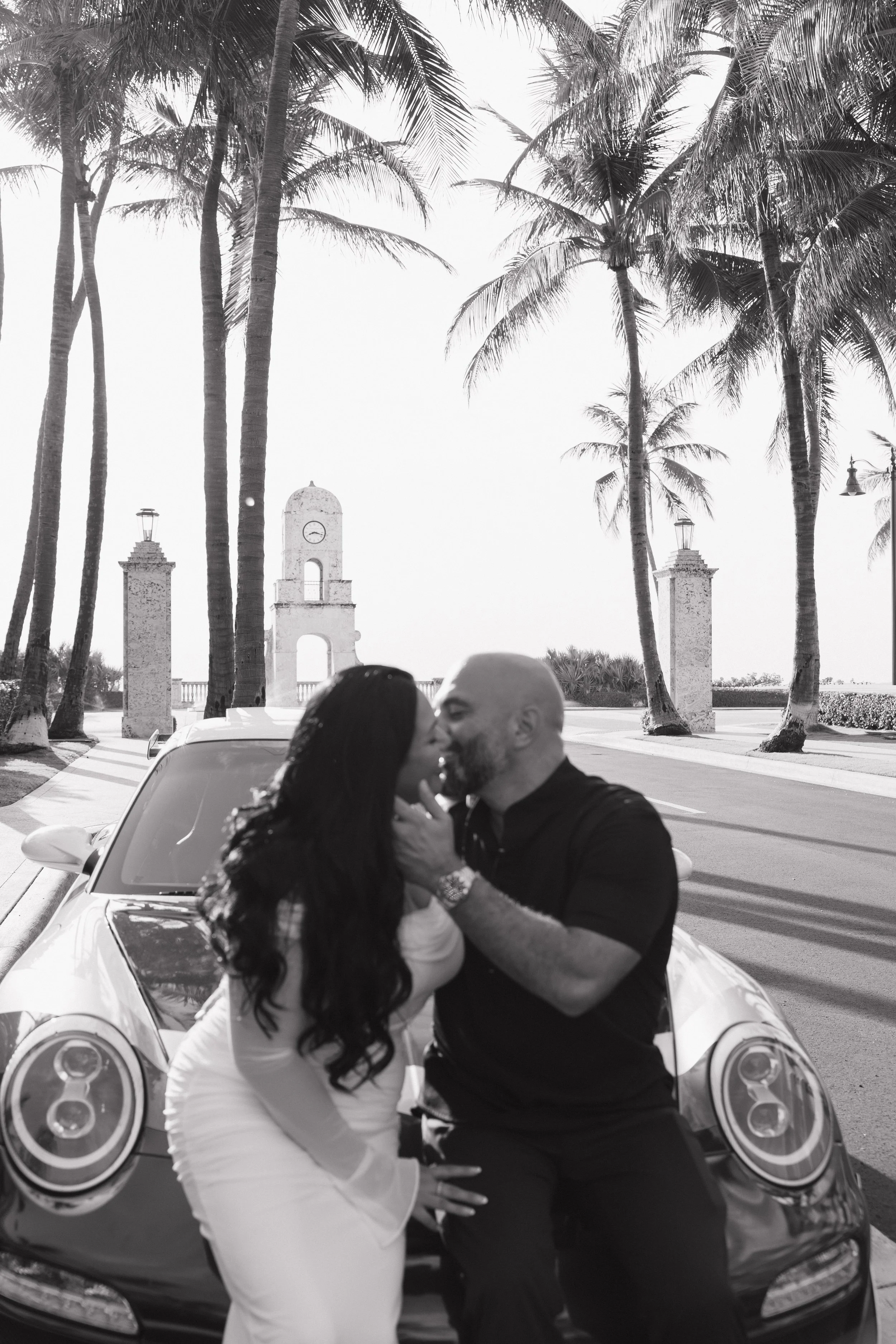 A black and white photo of a couple kissing in front of a car, with palm trees and historical structure in the background.