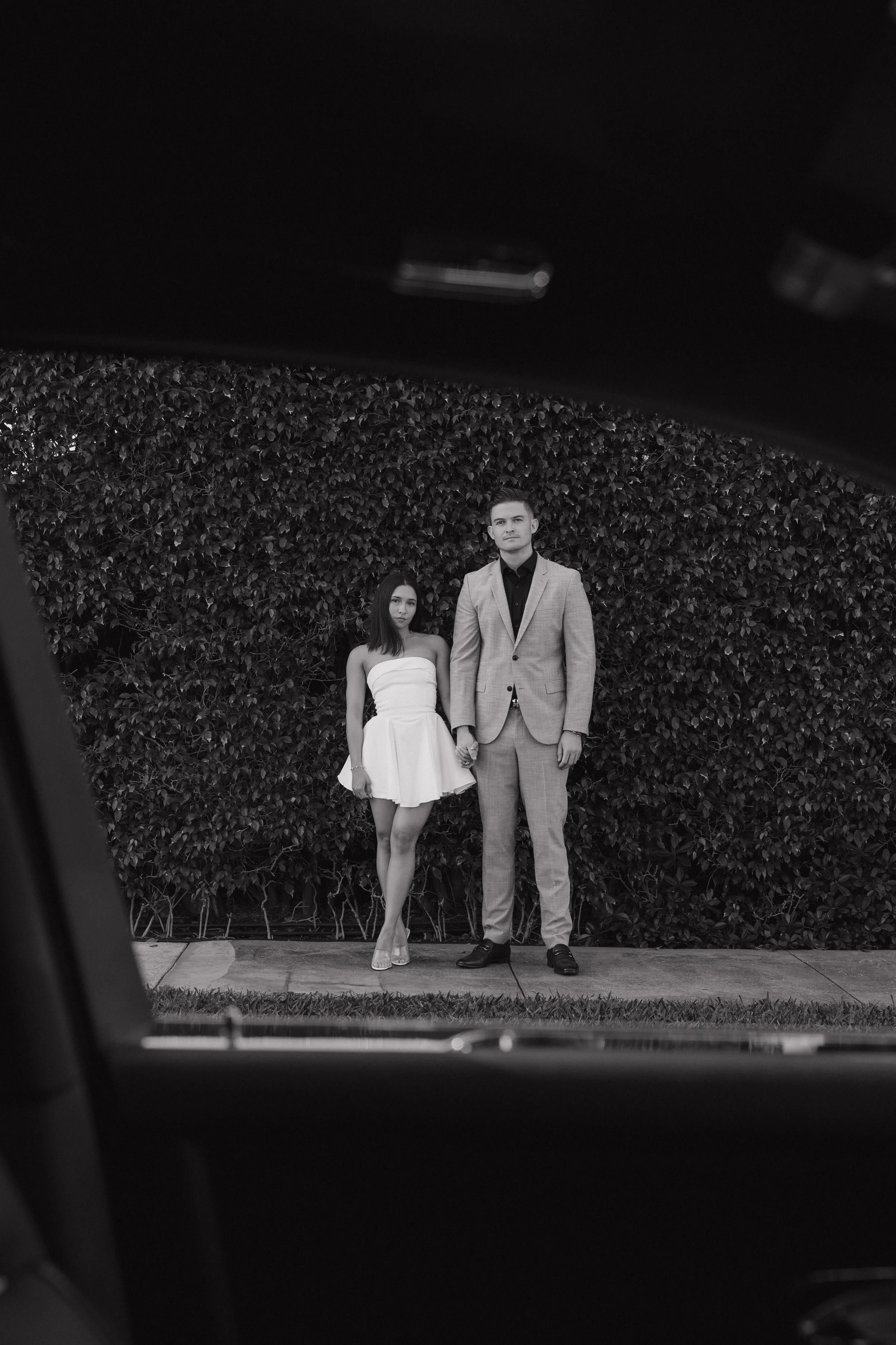 A man and woman standing hand-in-hand in front of a hedge, viewed through a partially opened car door.