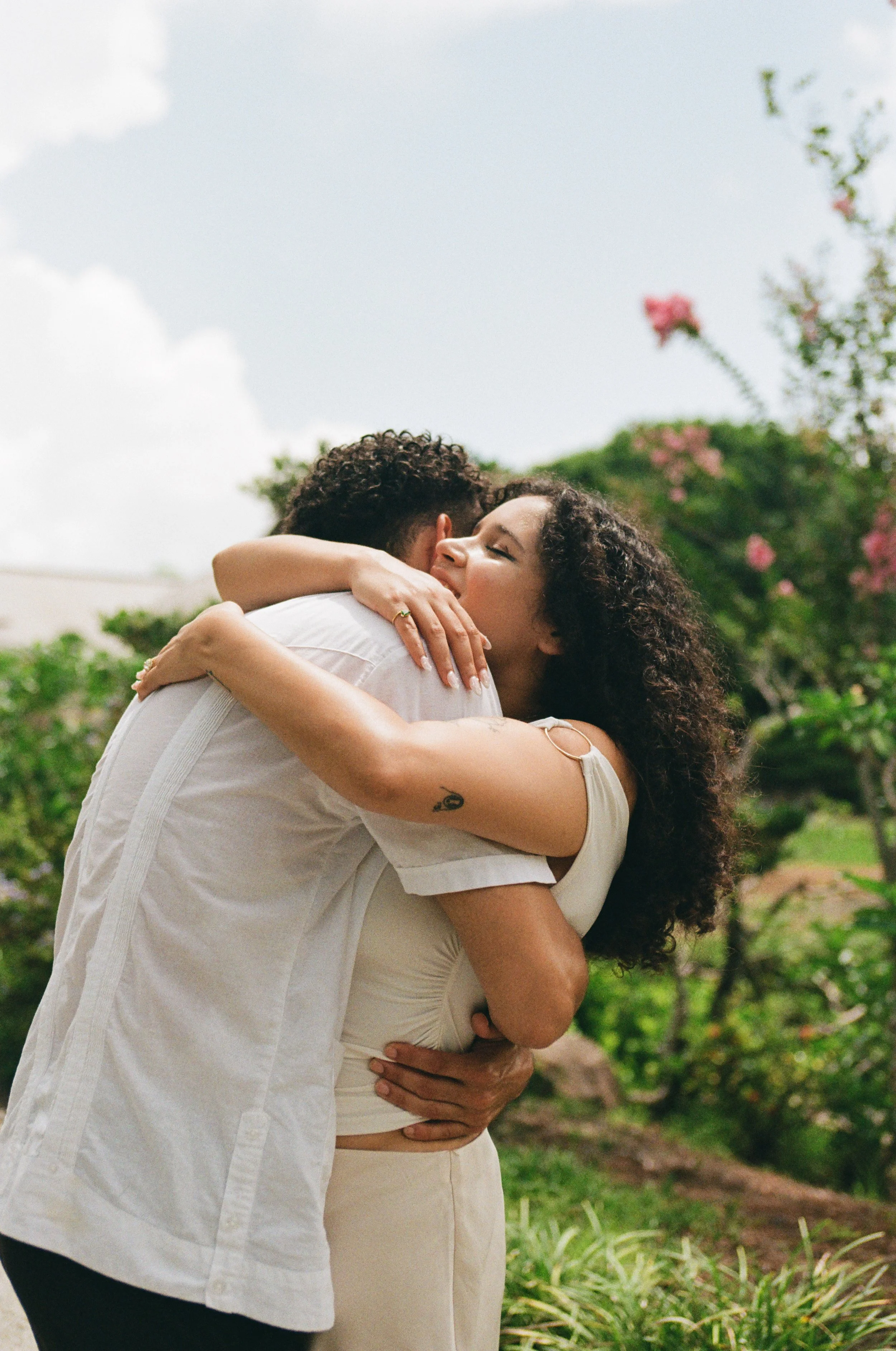 A woman and a man embrace outdoors in a garden with pink flowers and green foliage, with the woman smiling and eyes closed.