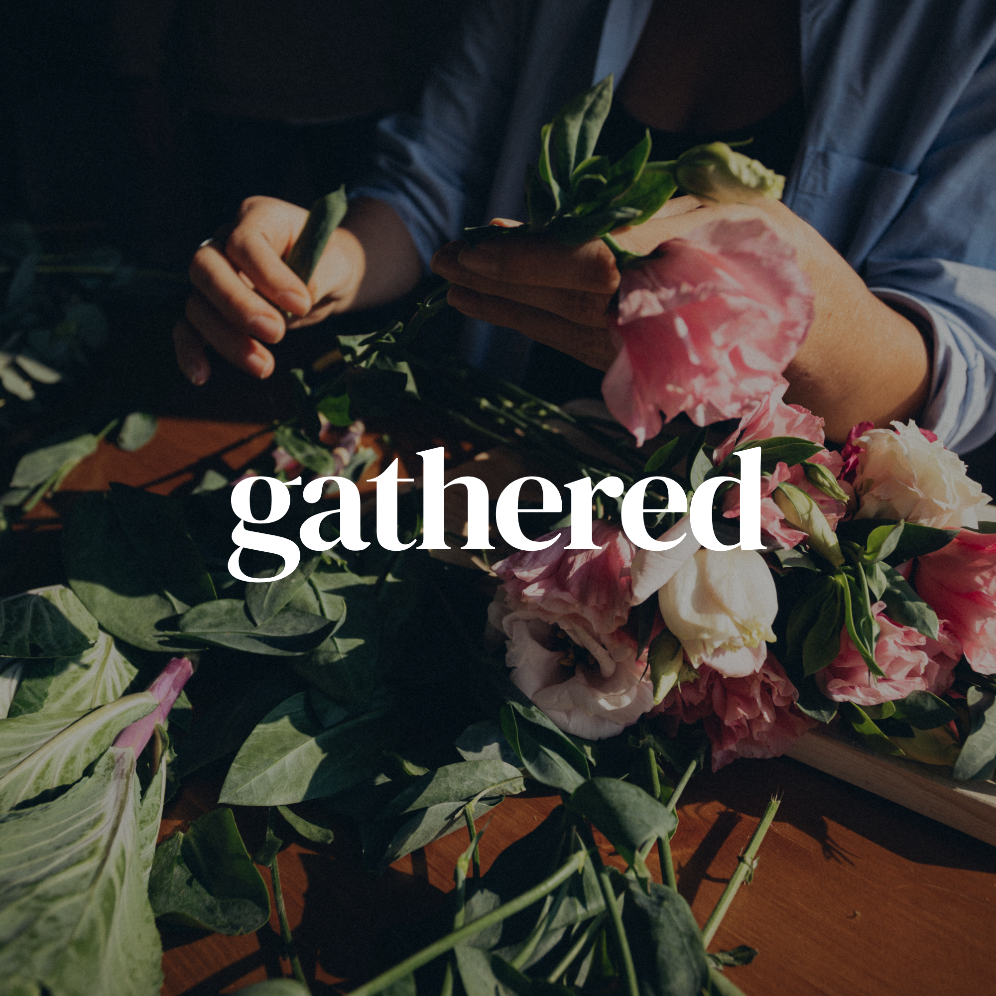 A person arranging a bouquet of pink and white flowers on a wooden table, wearing a blue jacket and a black shirt.