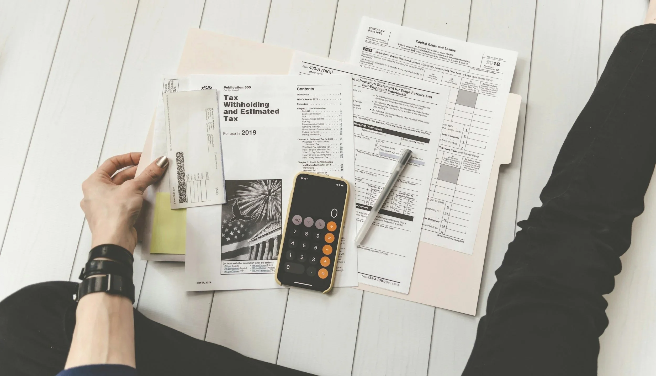 Person working at a white table with tax forms, a calculator, a pen, and money, indicating tax preparation activity.