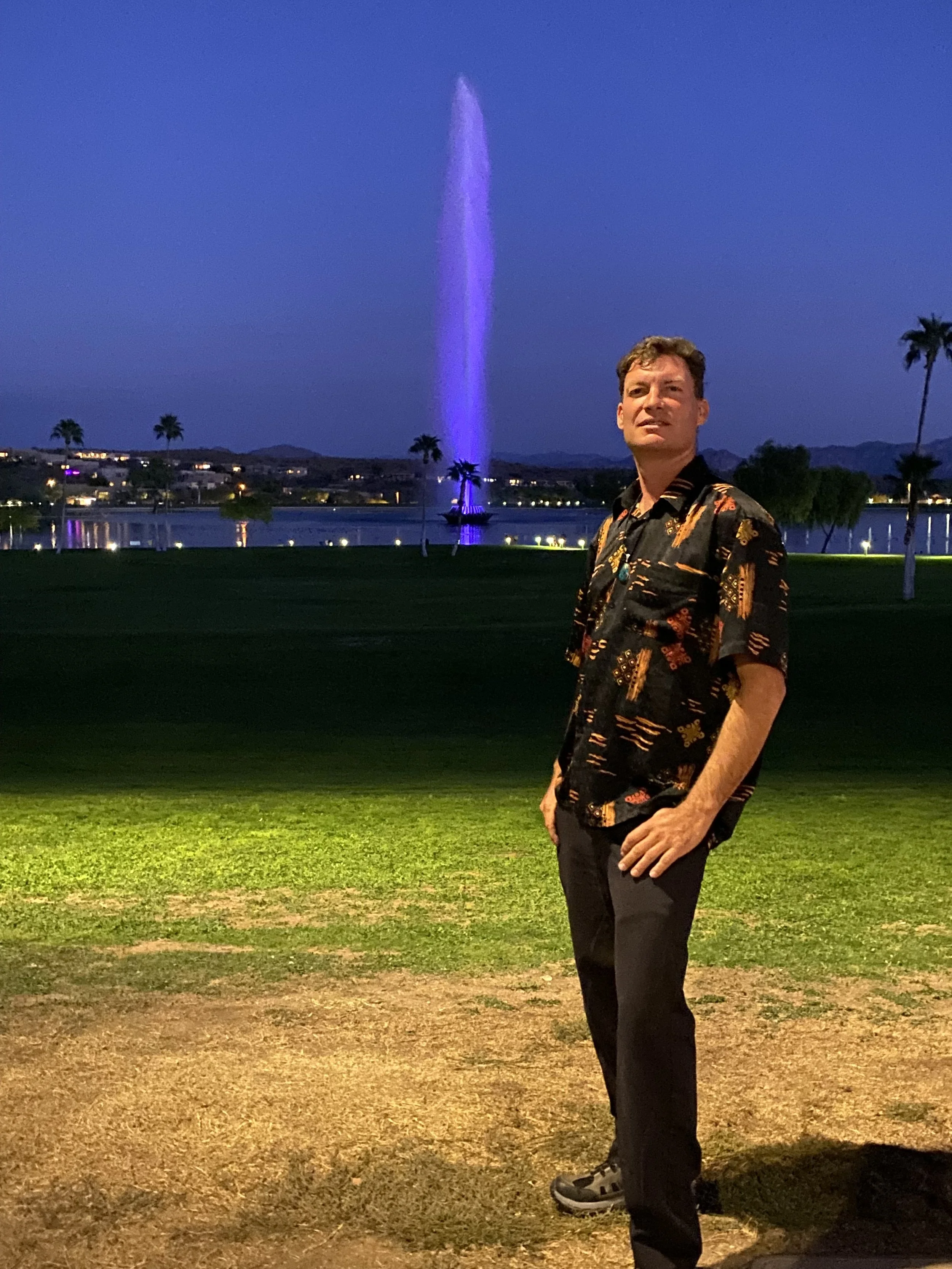A man stands on a grassy area at night, with a body of water and a fountain illuminated in purple behind him. There are palm trees and distant mountains in the background.