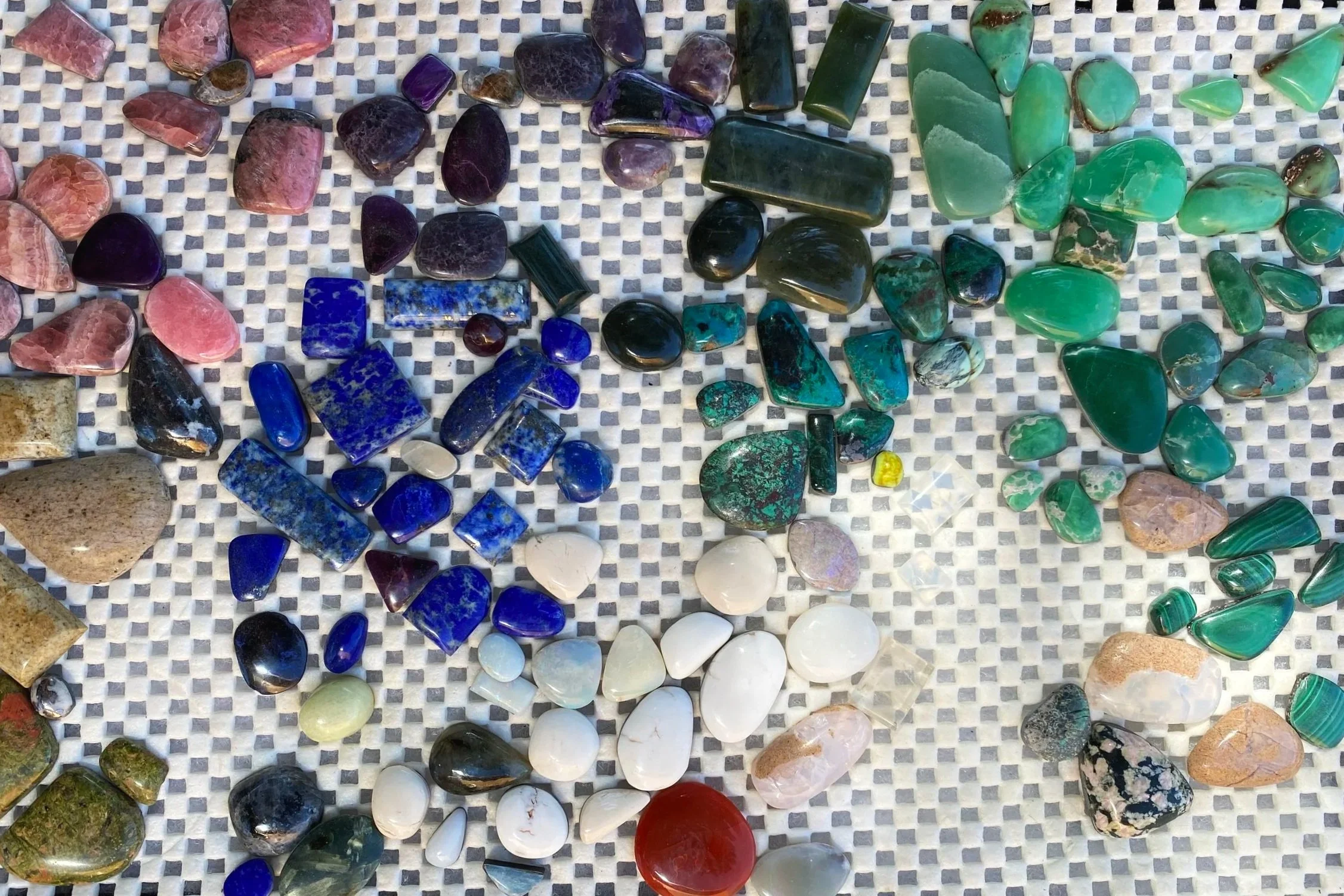 Various polished gemstones and crystals in different colors including pink, purple, blue, green, black, white, and red, arranged on a gray and white checkered surface.