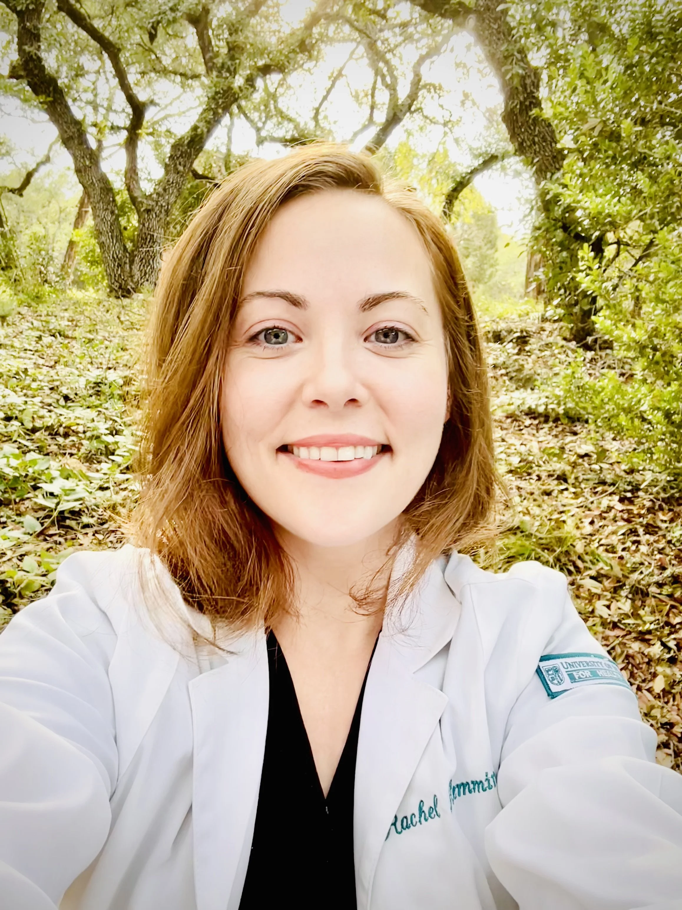 A woman with red hair smiling and taking a selfie outdoors. She is wearing a white doctor coat with a patch that reads 'Rachel' on it, and there are trees and fallen leaves in the background.