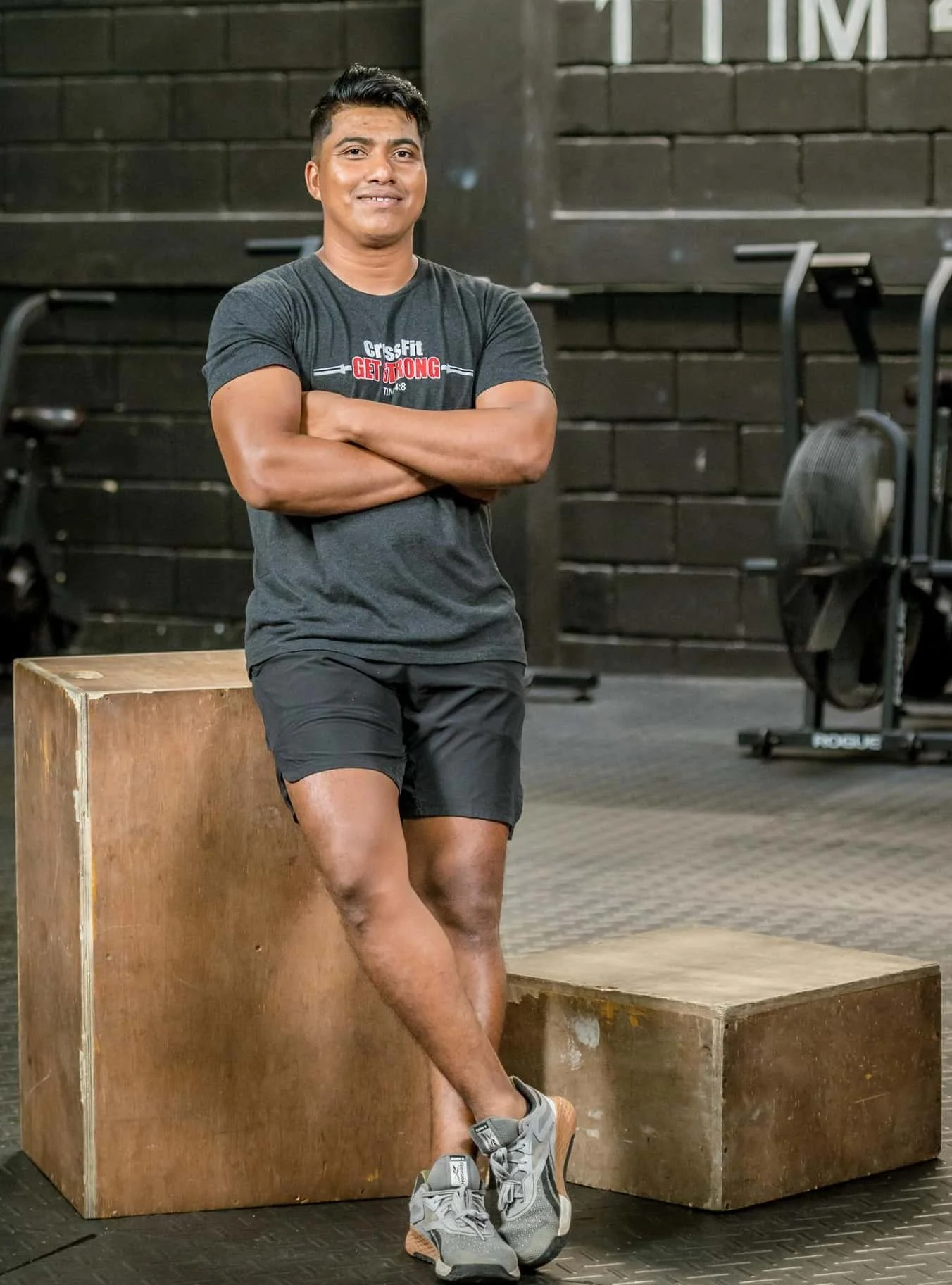 A man standing with arms crossed and leaning against a wooden box in a gym. He is wearing a dark grey t-shirt, black shorts, and grey running shoes. The background shows gym equipment and a black wall with large white lettering.