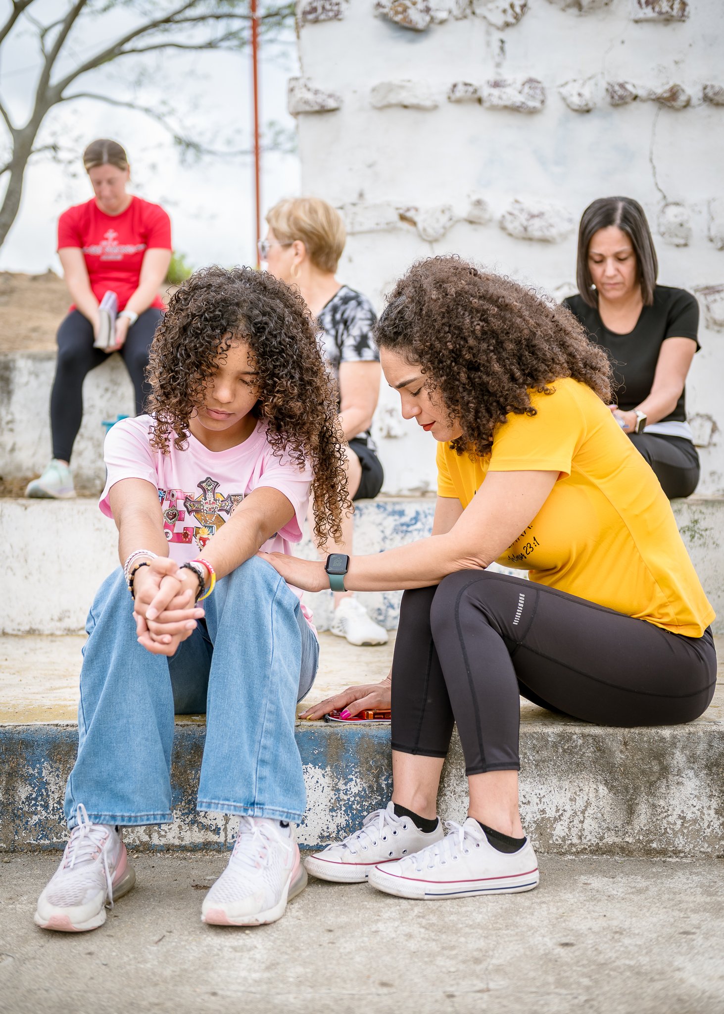 Two young women sit on a step, holding hands and looking down. Two women sit behind them, looking down or away, against a stone wall outdoors. The scene suggests a moment of reflection or comfort.