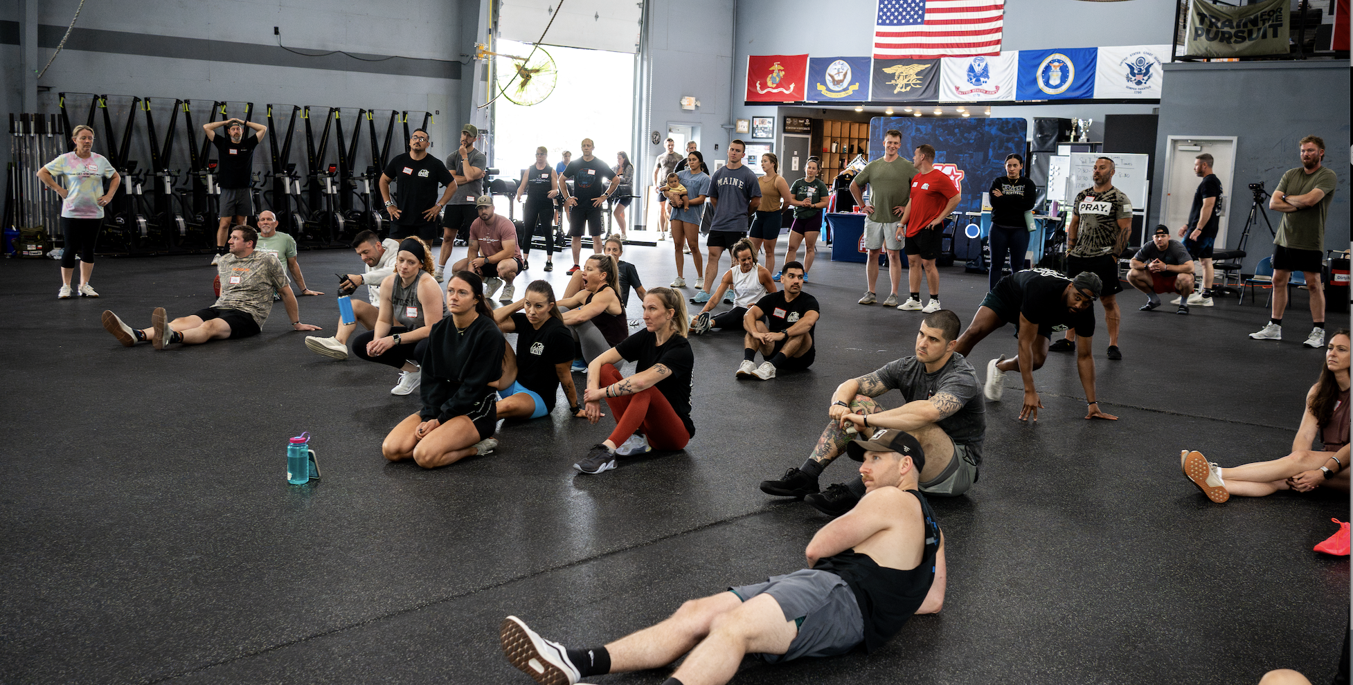 A large group of people gathered inside a gym, sitting and standing, with some on the floor and others near equipment, watching an event or presentation. The gym has flags and banners hanging on the walls.