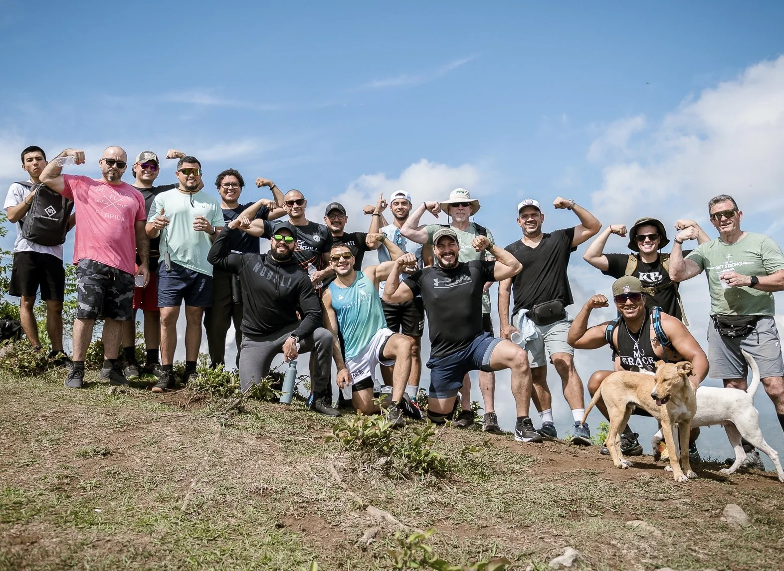 Group of people outdoors on a hilltop, flexing their muscles and smiling, with dogs at their feet, under a partly cloudy sky.