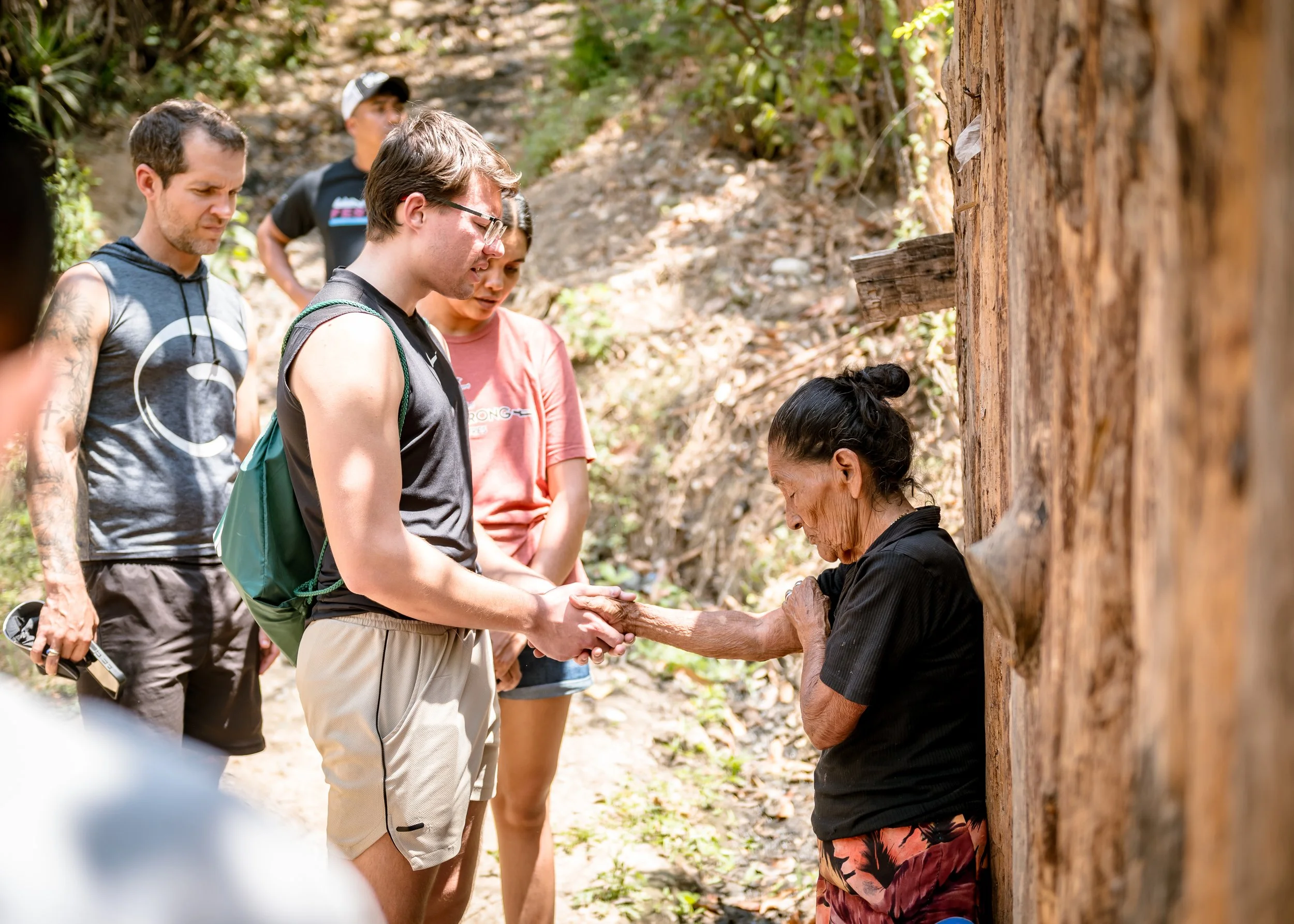 A group of young adults standing outdoors in a wooded area, attentively listening to an elderly woman who is holding hands with one of the young men, near a wooden structure.