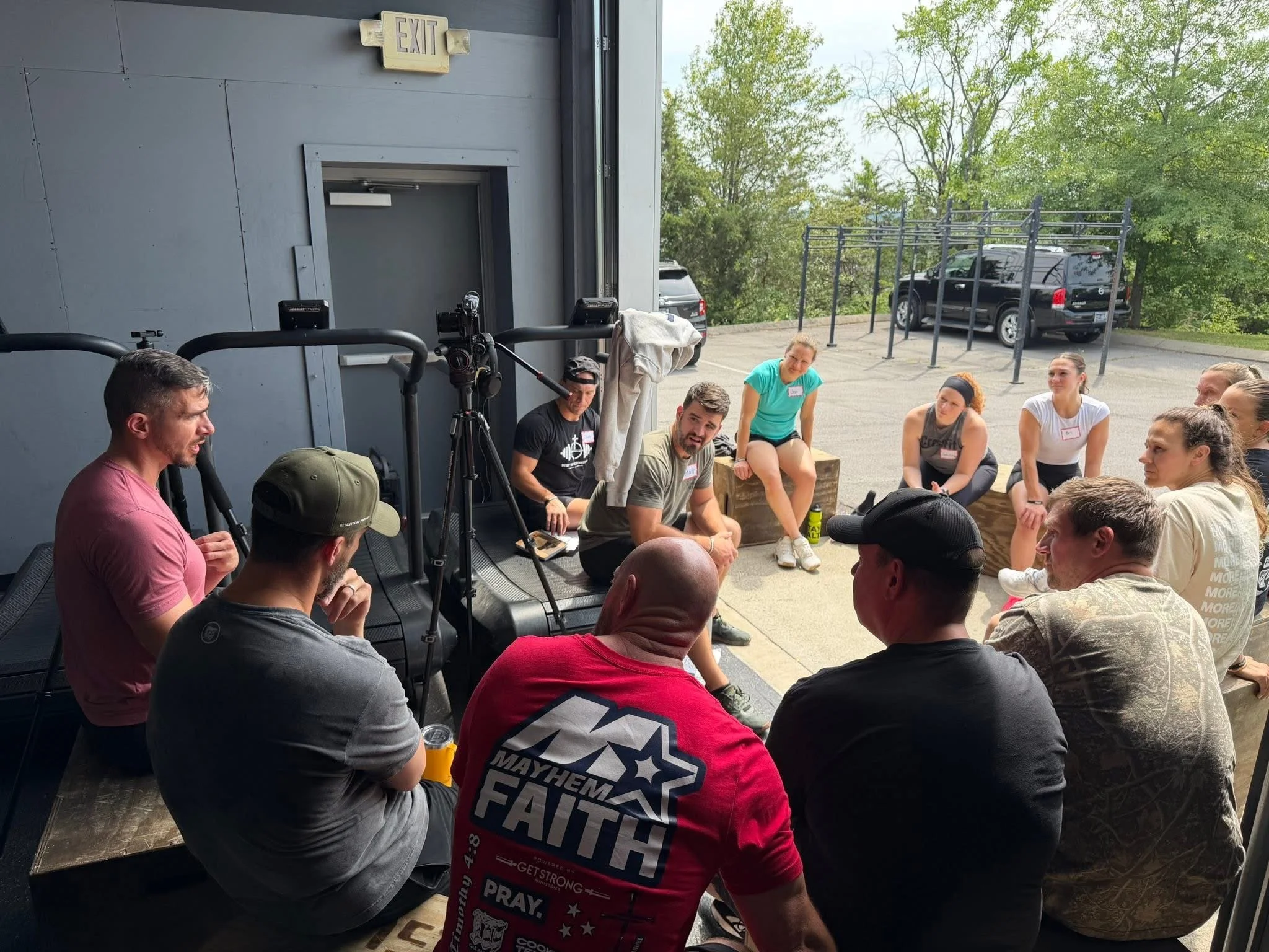 A group of people sitting outdoors in a circle, engaged in conversation. The setting appears to be a workout or fitness event, with a camera and some equipment visible nearby. Trees and parked cars are in the background.