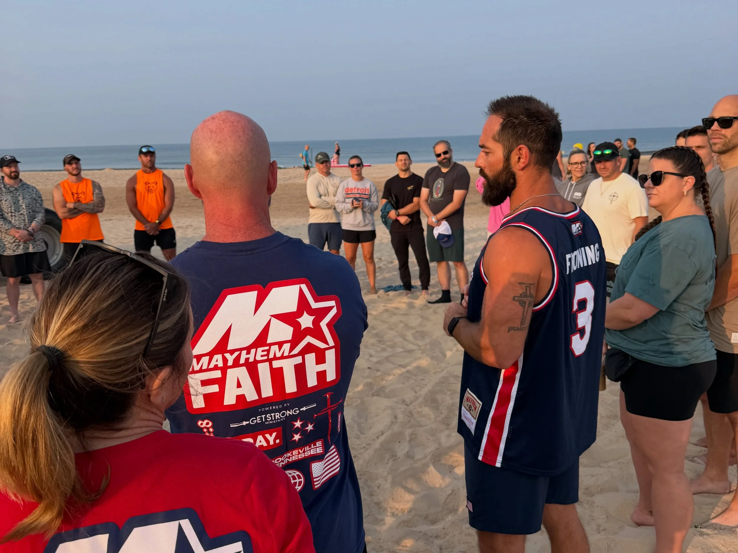 Group of people gathered on a beach, some wearing sunglasses and casual clothes, listening to a man with a beard and a basketball uniform speak. The ocean and a clear sky are visible in the background.