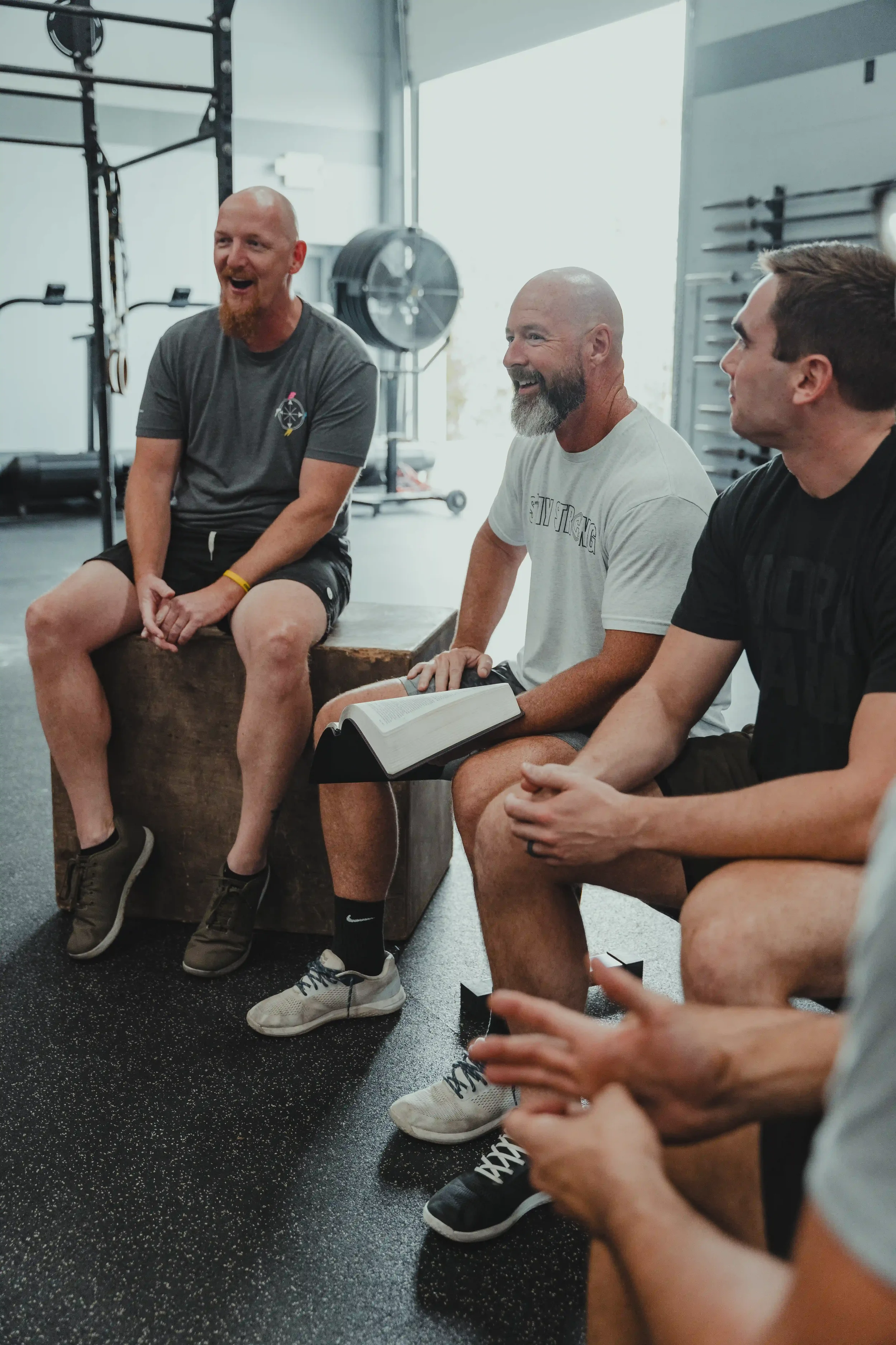 A group of men sitting in a gym, engaged in conversation and laughter, with gym equipment in the background.