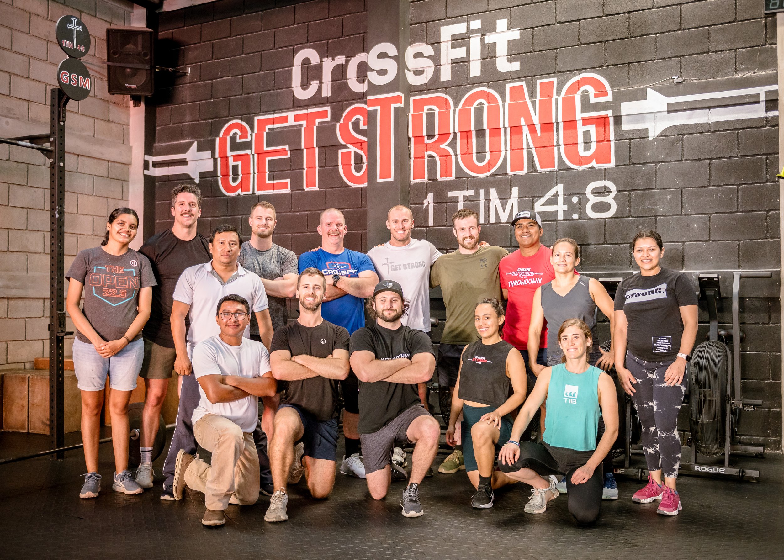 Group of people posing inside a CrossFit gym with a large sign on the wall reading "CrossFit GET STRONG" and a timer showing 4:48.