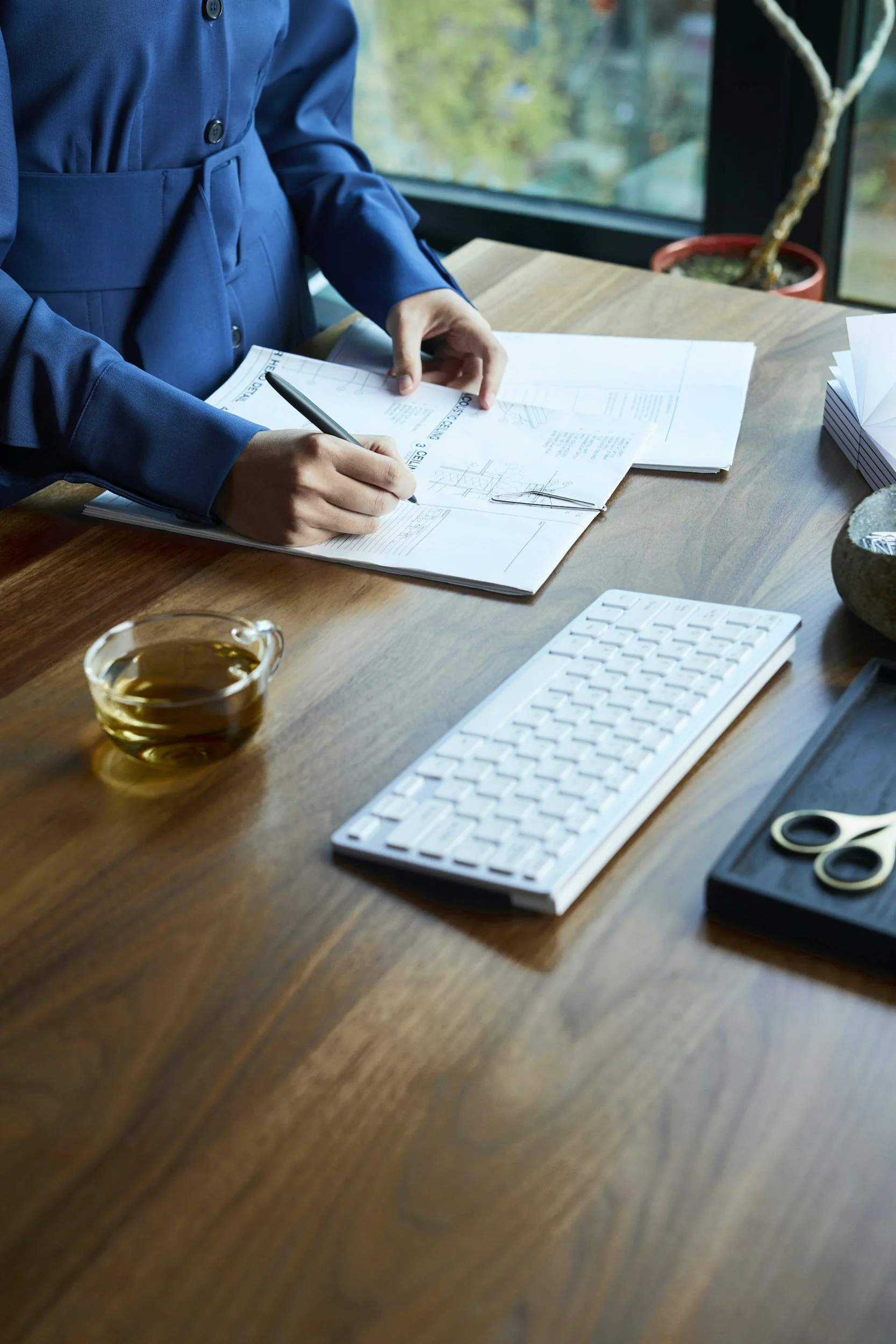 Person in a blue blazer sitting at a wooden desk working with papers, a pen, and a clipboard. Items on the desk include a glass of tea, a white keyboard, a pair of scissors, and a black tray with audio headsets. A window with a view of greenery and a potted plant is in the background.