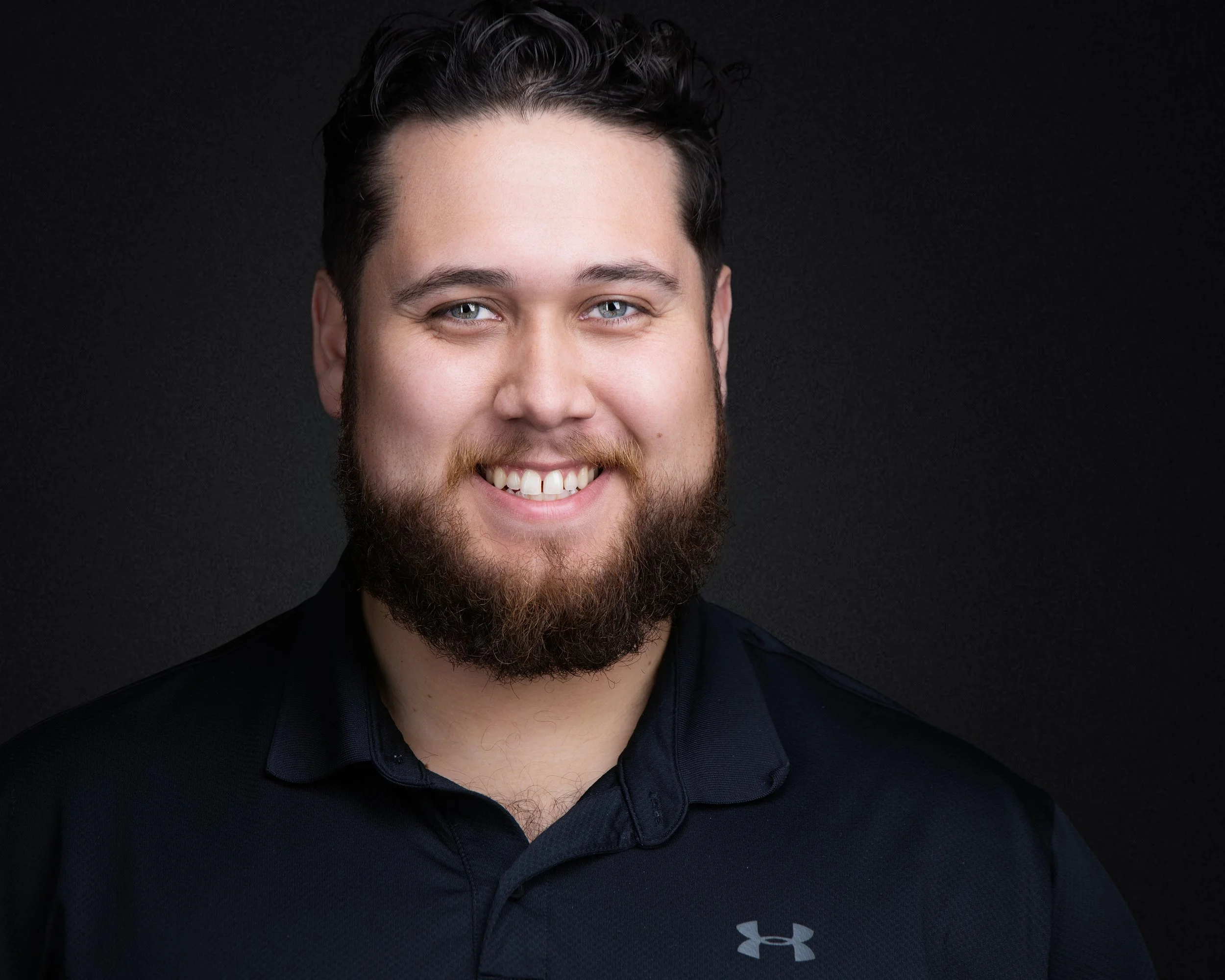 Close-up portrait of a smiling man with light skin, dark curly hair, blue eyes, and a beard, wearing a black sports shirt with the Under Armour logo, against a black background.