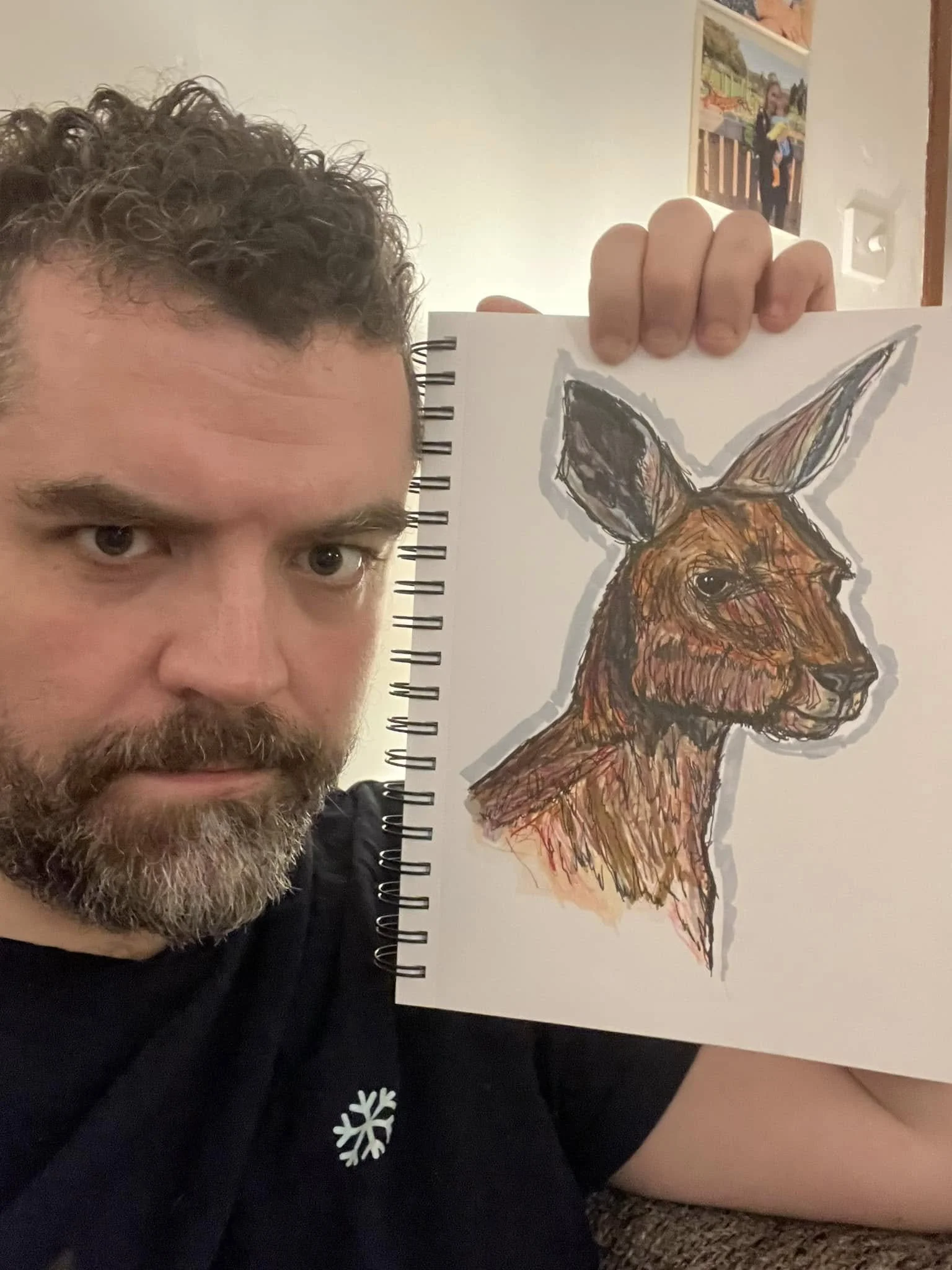 A man with curly hair holding a spiral-bound sketchbook with a colorful drawing of a fox on it, partially covering his face.