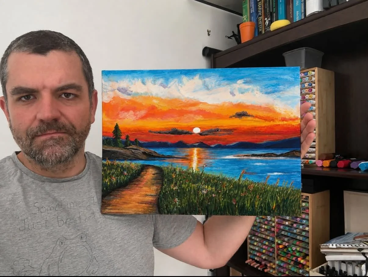 A man holding a colorful painting of a sunset over a lake with mountains in the background, a path and grassy foreground, while standing in a room with art supplies and bookshelves.