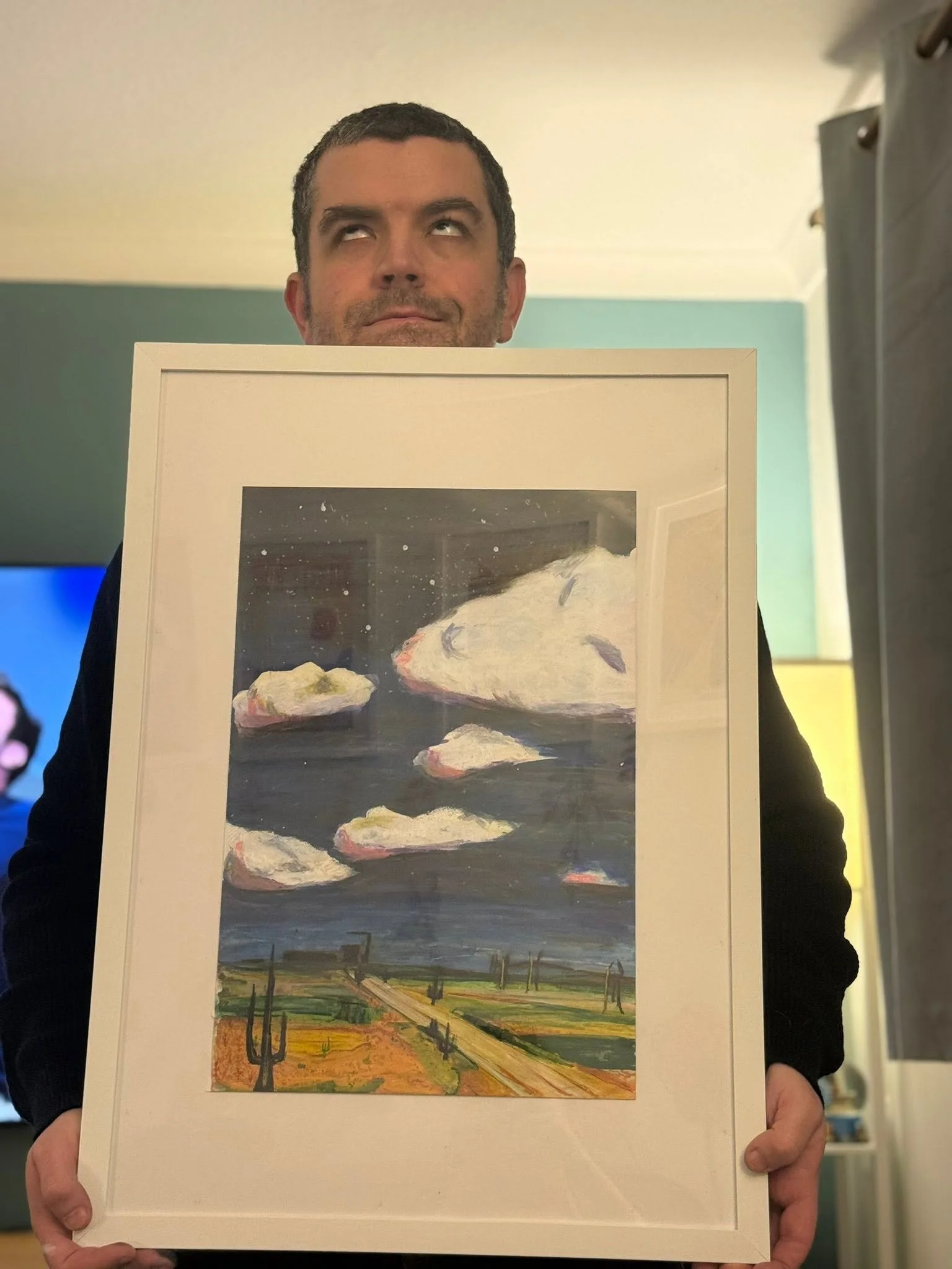 Person holding a framed landscape painting with clouds in the sky and a field with trees and a dirt road.