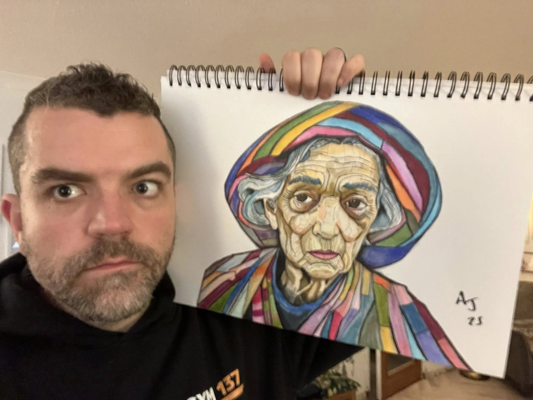 A person with short curly hair and a beard taking a selfie with a colorful portrait of an elderly woman with a rainbow-colored headscarf behind them.