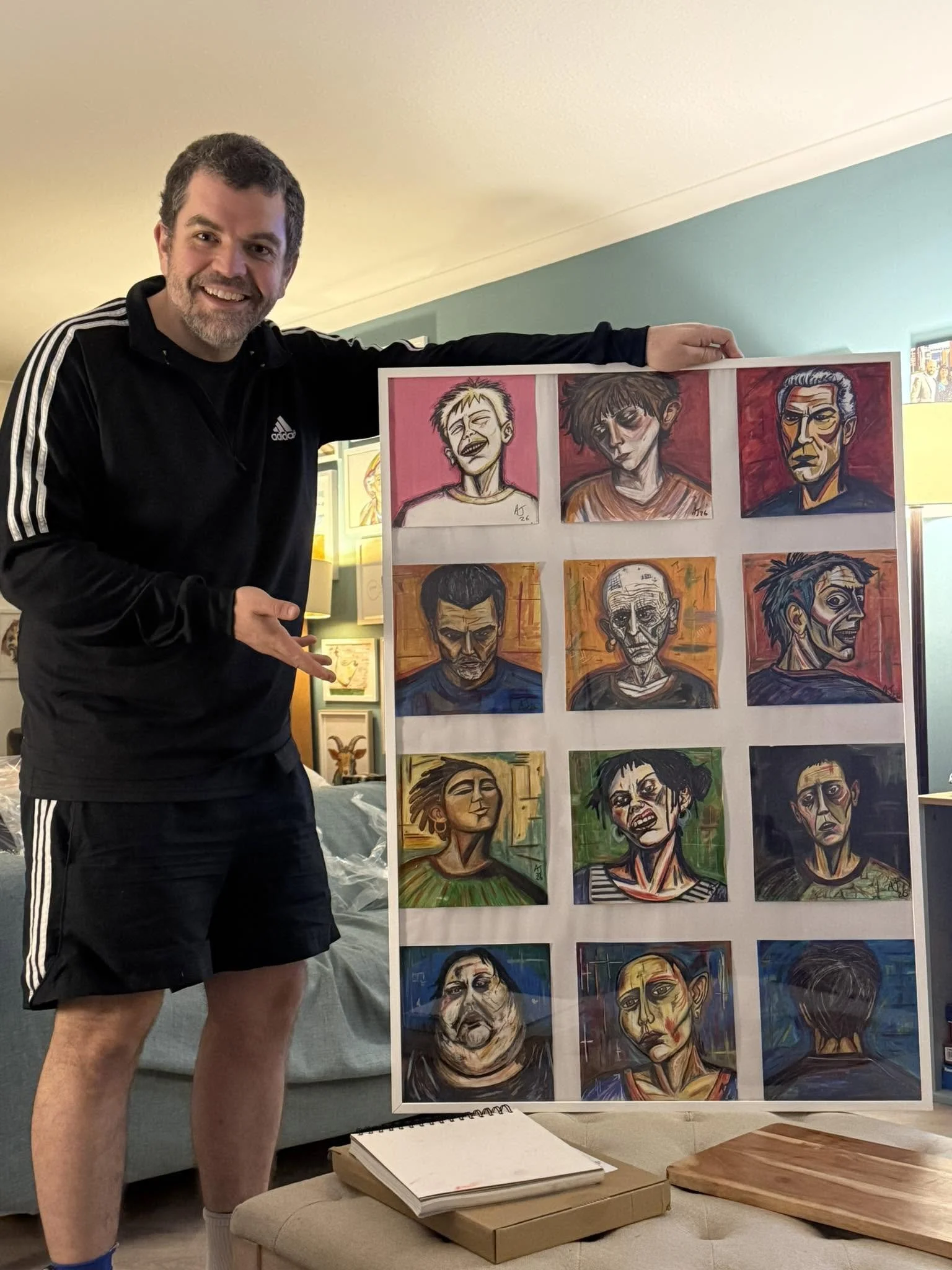 A smiling man in a black Adidas jacket and shorts points to a display of colorful portrait artwork. The artwork features expressive faces painted in a variety of styles. The setting appears to be an art gallery or studio with other framed pieces visi