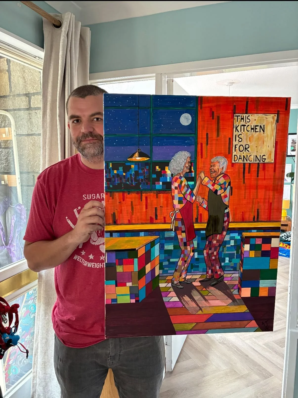 A man holding a colorful painting of two elderly women dancing in a kitchen, with a cityscape and moon visible through the window in the background.