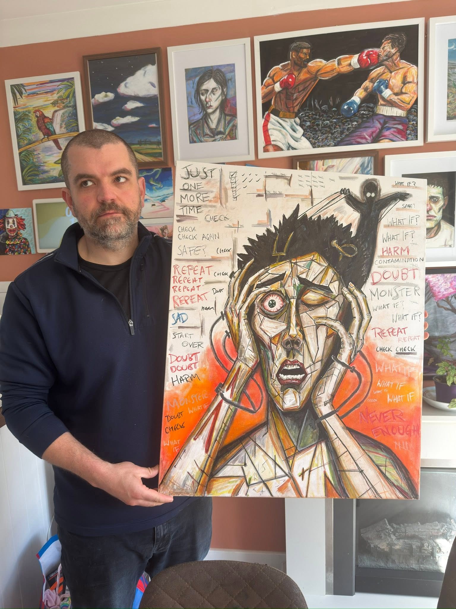 A man with a beard and wearing a black jacket is holding a colorful painting of a woman with a distressed expression, her hands on her head, surrounded by words and phrases like "Doubt," "Repetition," and "What if?". Behind him, there are several framed artworks on a wall, including paintings of a boxer fight, a portrait, and landscapes.
