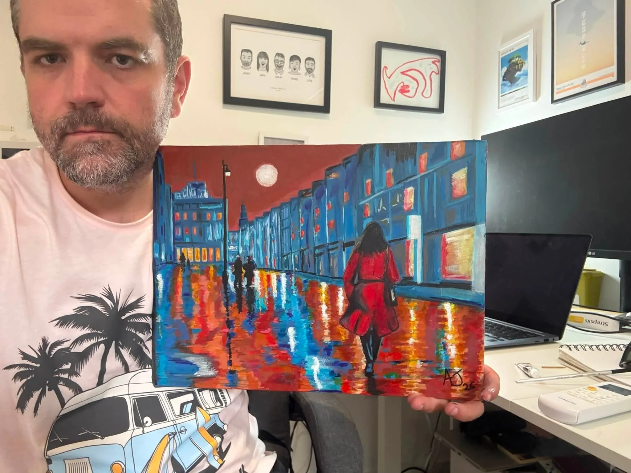 Man holding a painting of a city street scene at night with colorful reflections on wet pavement and a woman in a red coat walking away.