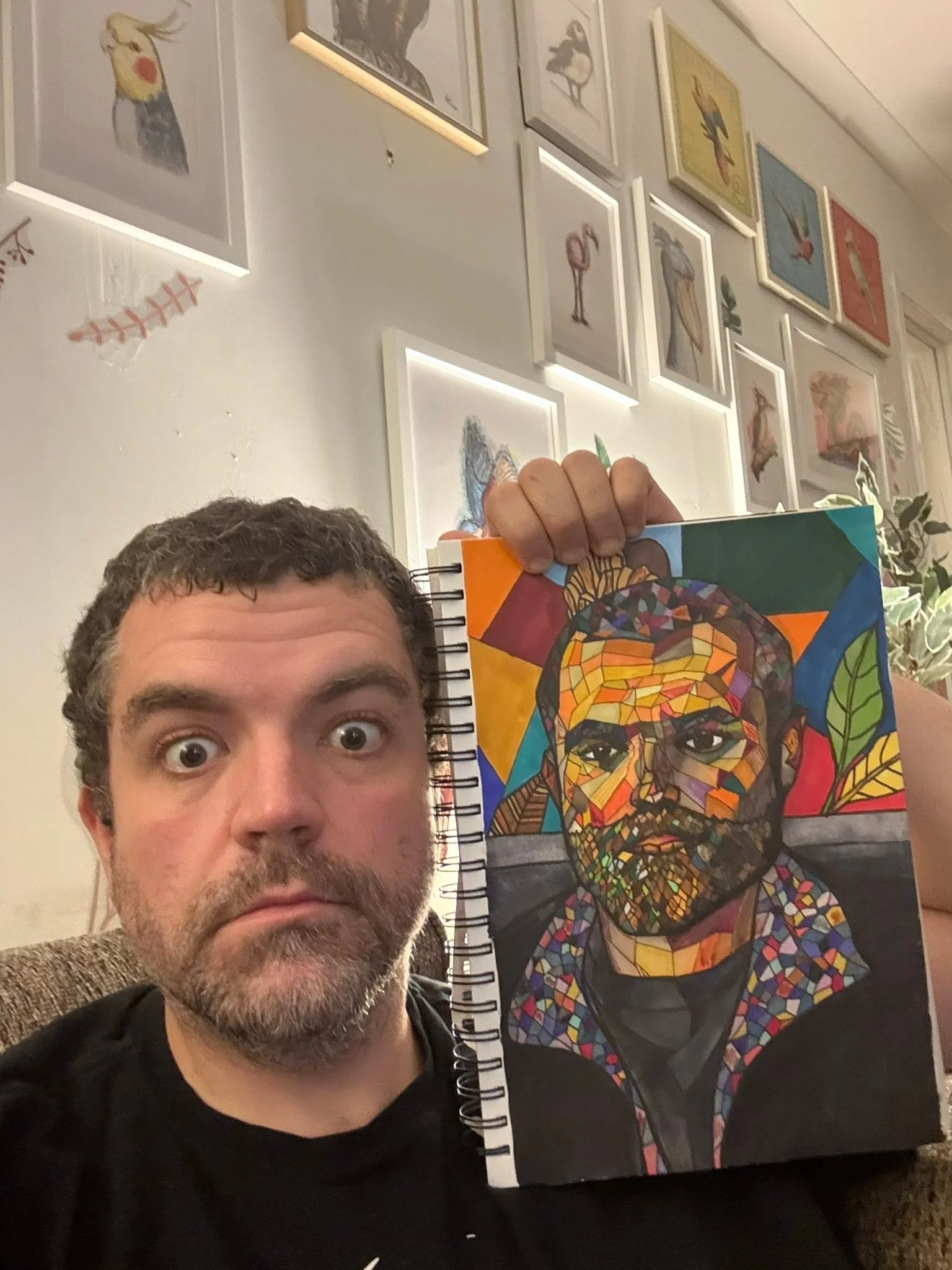 A man with curly brown hair, a beard, and blue eyes is taking a selfie in front of a wall decorated with art. He is holding a colorful canvas featuring a mosaic-style portrait of a person's face. The wall has various framed pictures, mostly of birds,