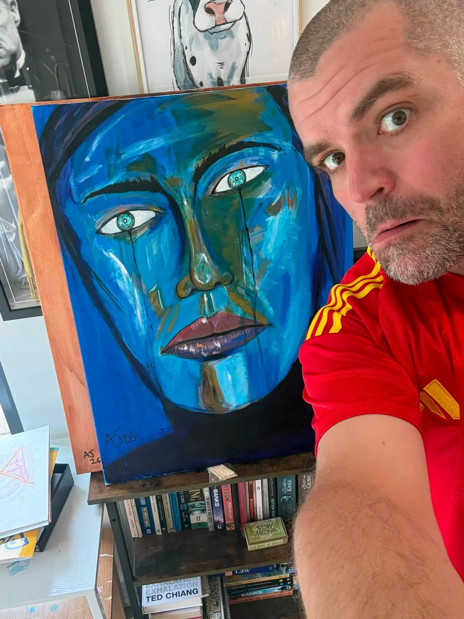 A man with short hair and a beard taking a selfie next to a colorful painting of a human face with blue and green tones, on a wooden easel. The background includes a white wall with artwork and a bookshelf.