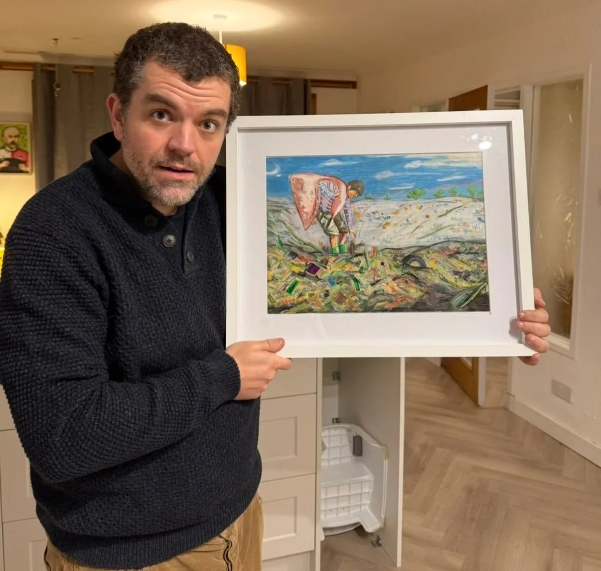 A man with dark hair and a beard holding a framed colorful abstract painting of a person in a landscape with a blue sky, green trees, and a patterned ground.