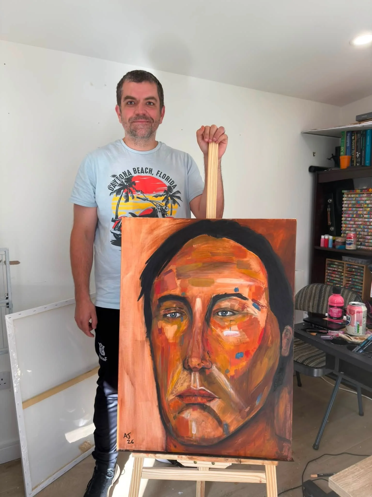 A man standing next to an abstract portrait painting of a woman's face on an easel in an art studio. The man is wearing a light T-shirt with a graphic of a motorcycle and palm trees that says "Daytona Beach, Florida." The studio has books, art suppli
