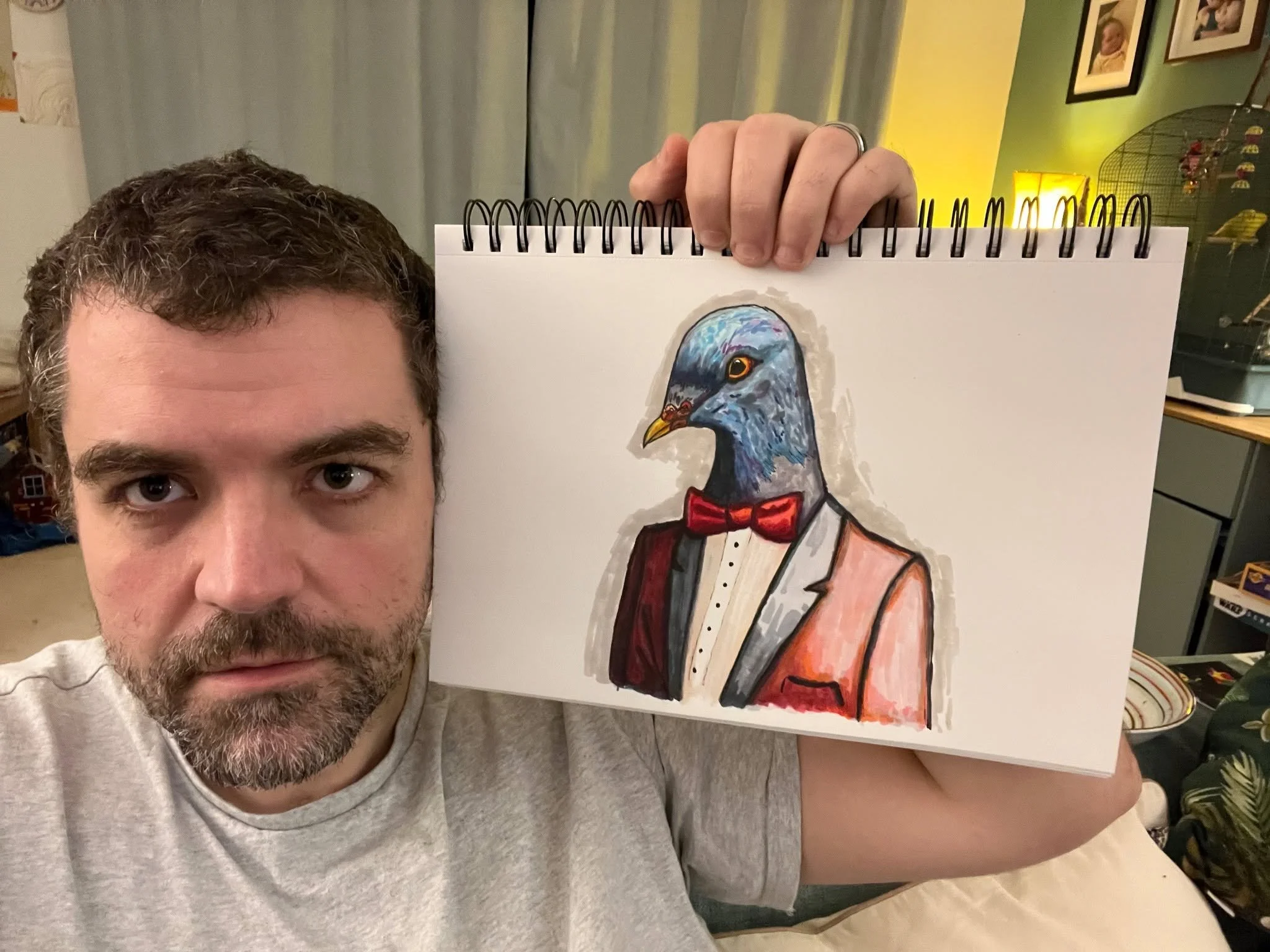A man with brown curly hair and a beard taking a selfie while holding up a painting of a pigeon dressed in a tuxedo with a red bow tie. The background includes a green wall with framed photographs, a lamp, and a birdcage.