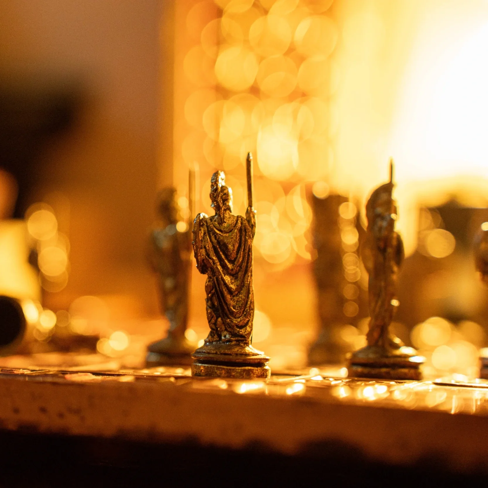 Close-up of a small ornate metallic statue of a religious figure, likely a saint, with a background of blurred warm lights.