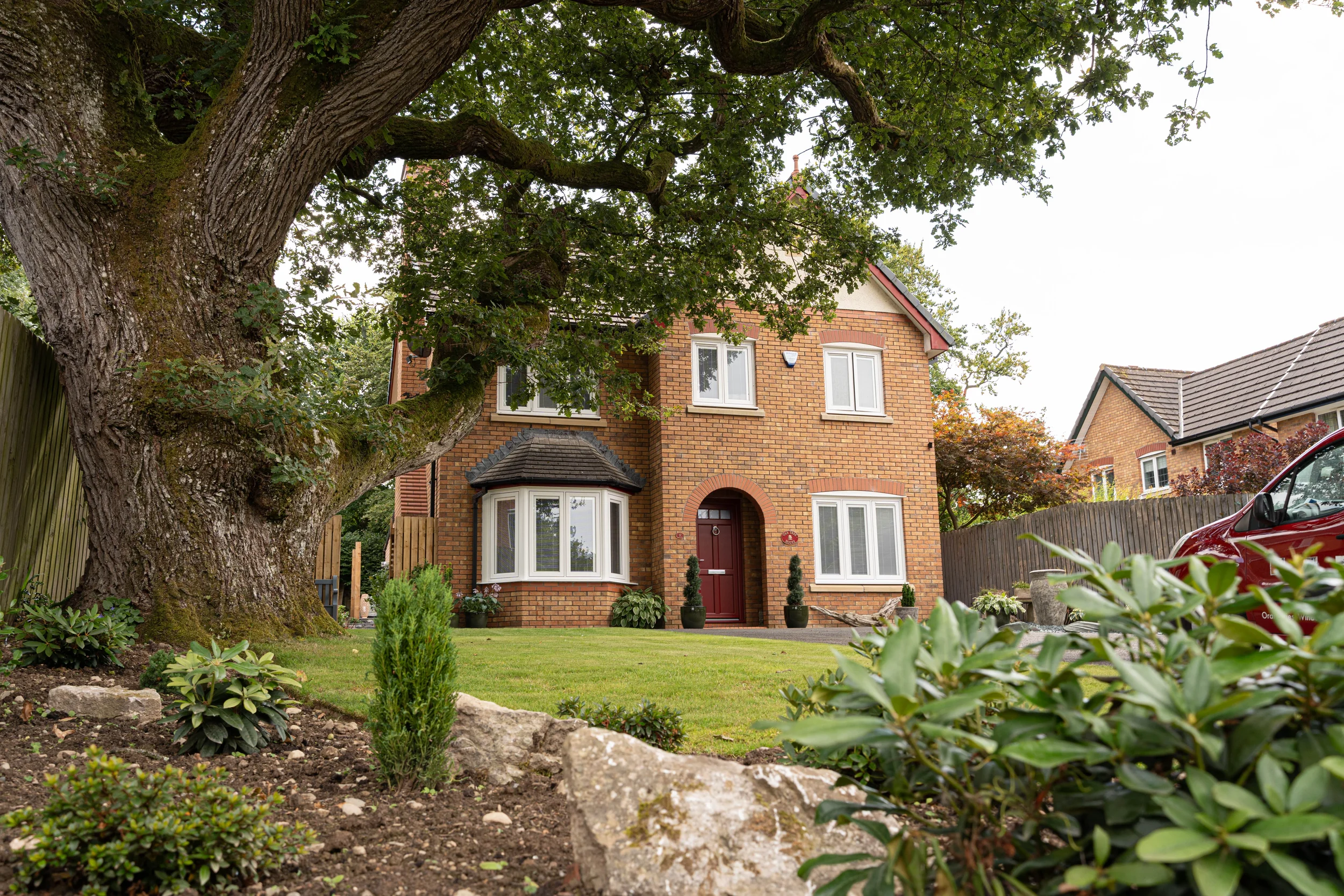 Front view of a two-story brick house with white-framed windows and a red front door, surrounded by a well-maintained garden with a large moss-covered tree, small bushes, and plants, with neighboring houses and a car partially visible.