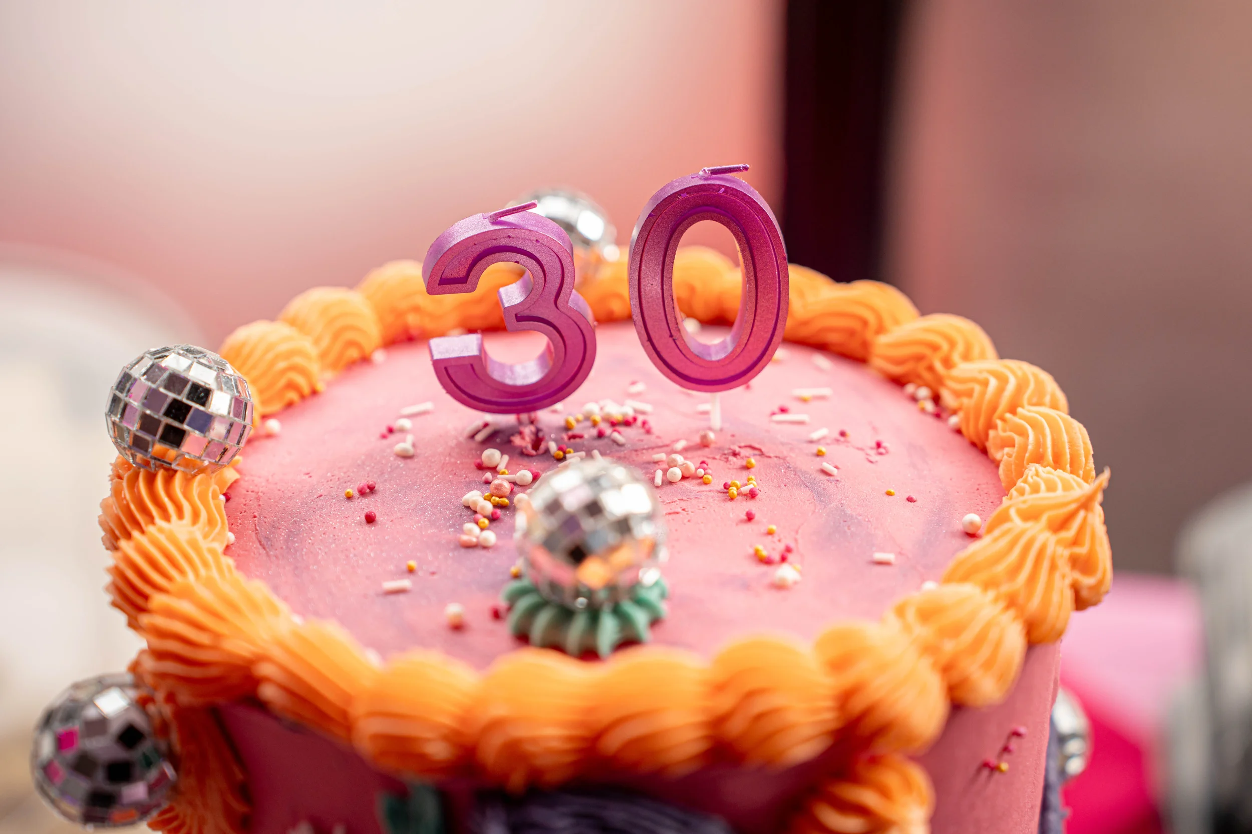A pink birthday cake decorated with orange frosting piping around the edges, colorful sprinkles, and silver disco ball cake toppers forming the number 30.