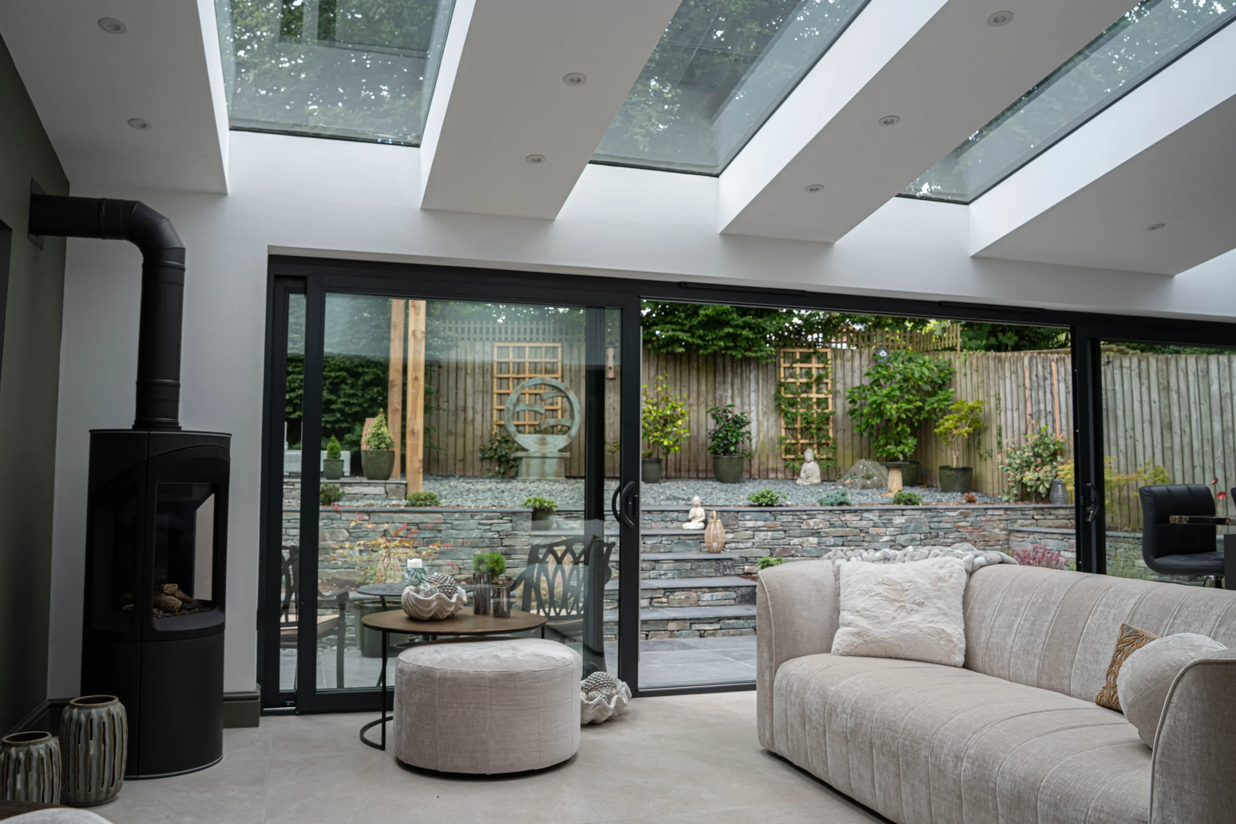A modern living room with a beige sectional sofa, a round ottoman, and decorative vases, with large glass sliding doors leading to a backyard garden with plants, a wooden fence, and garden statues.
