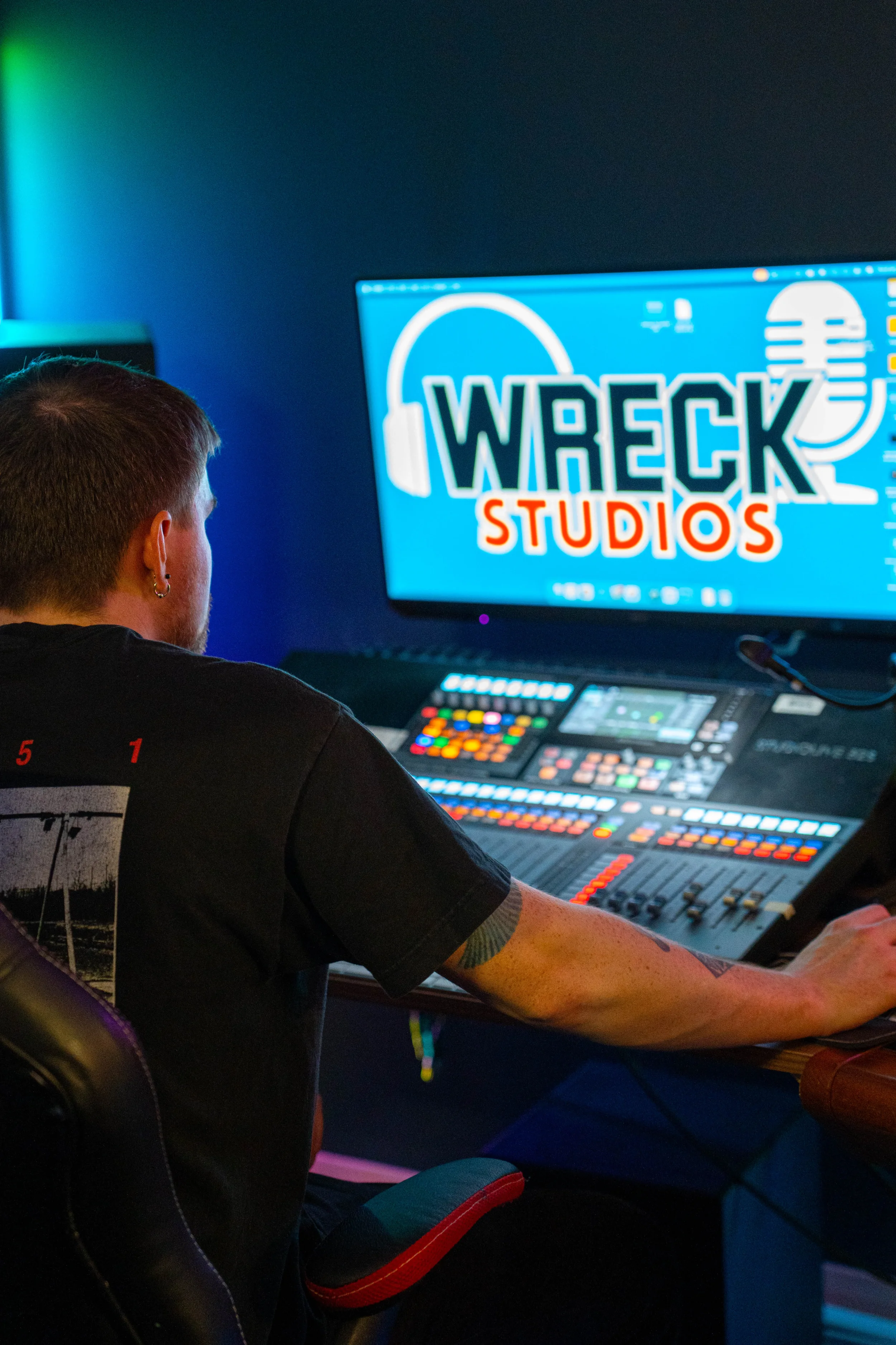 A person with a tattoo on their arm sitting at a mixing console in a studio, looking at a computer screen that displays a logo for WRECK Studios.