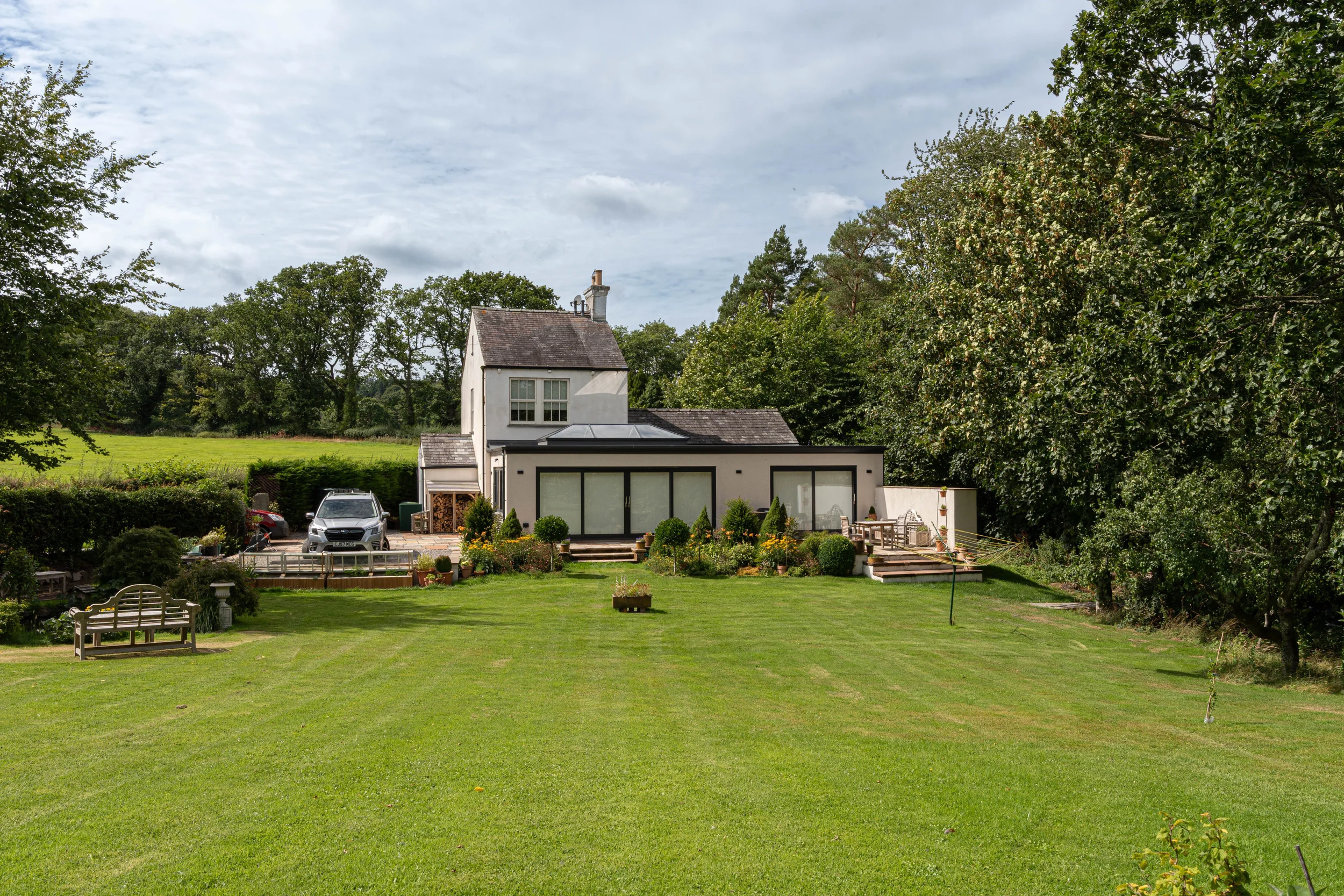A house with large glass doors, set in a lush green garden with trees and outdoor seating, under a partly cloudy sky.