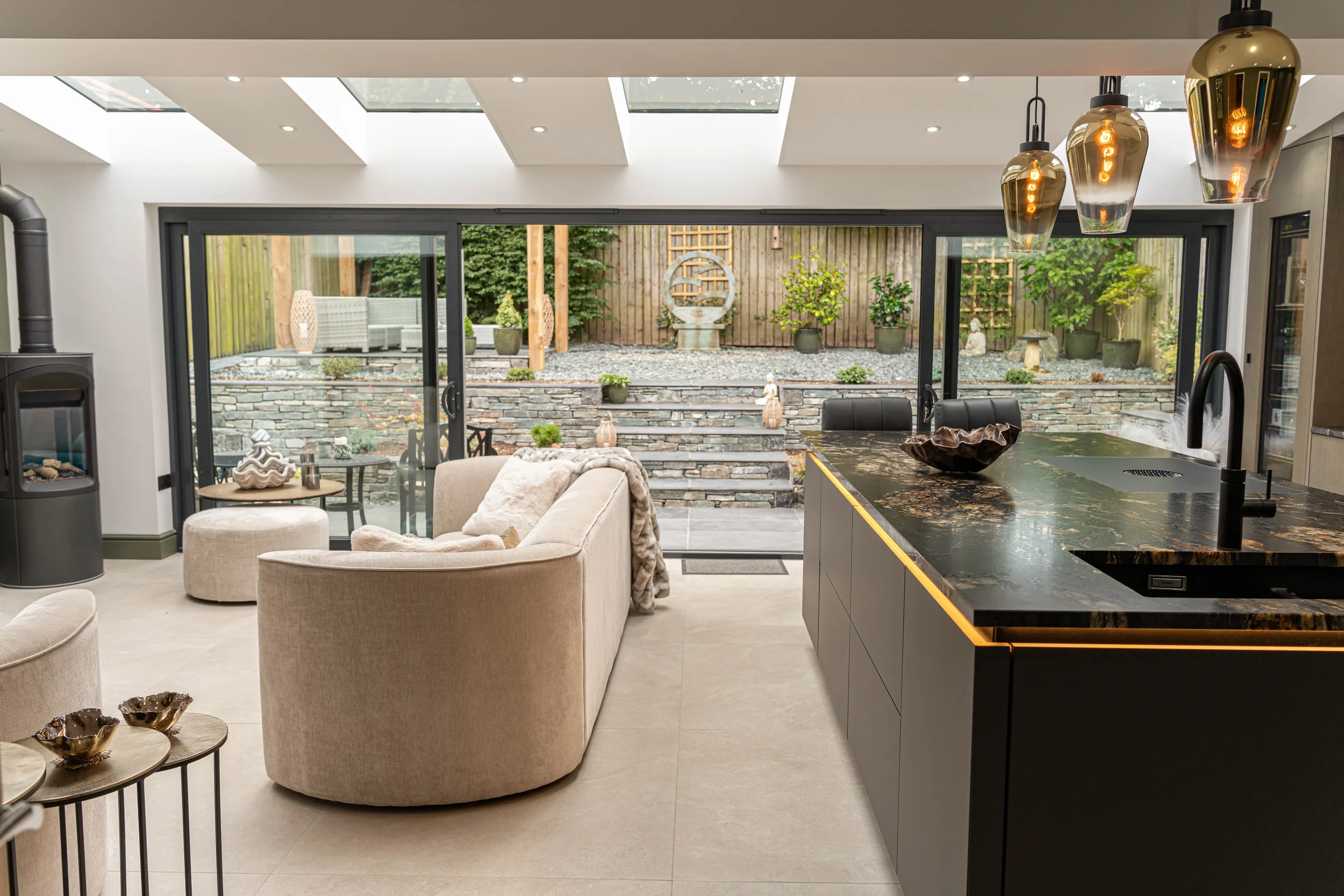 Modern living room with large glass sliding doors opening to a landscaped backyard with stone stairs, potted plants, and outdoor seating. Inside, there are beige armchairs, a black countertop with gold trim, pendant lights, and a stove.