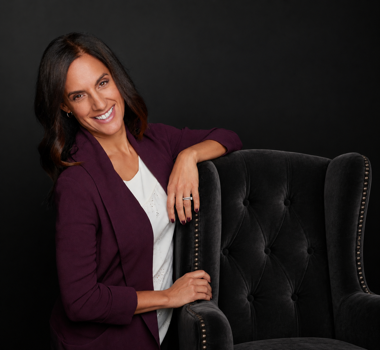A woman with dark brown hair styled in loose waves, smiling, wearing a white top and a burgundy blazer, resting her arm on the back of a black, tufted armchair against a solid black background.
