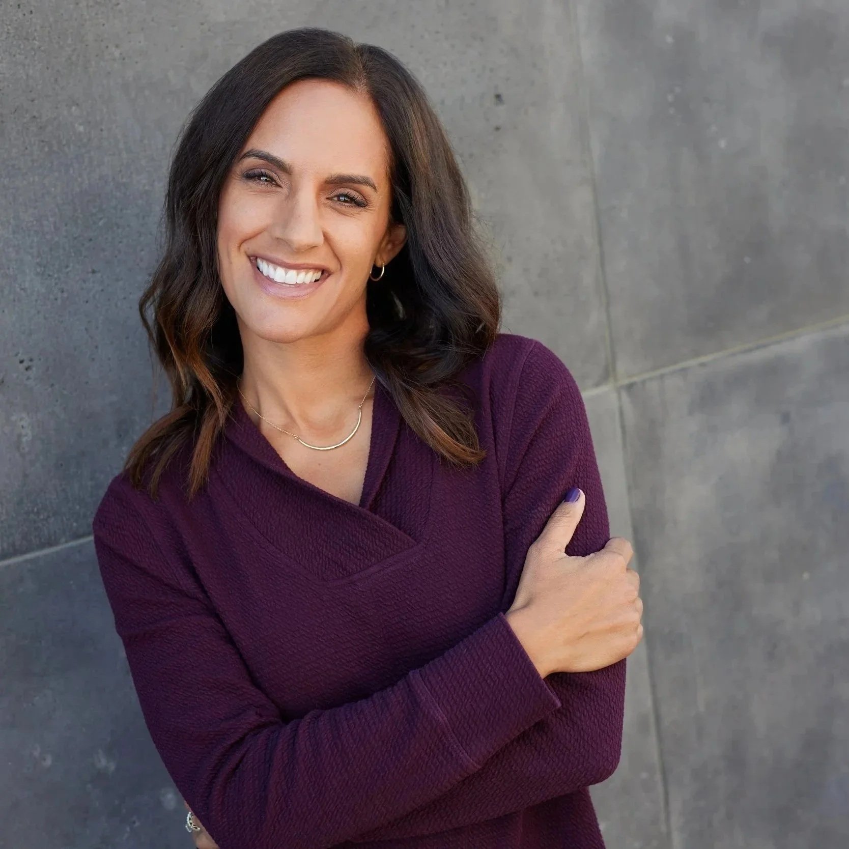 A woman with shoulder-length brown hair smiling and crossing her arms, wearing a purple sweater, standing against a plain gray wall.