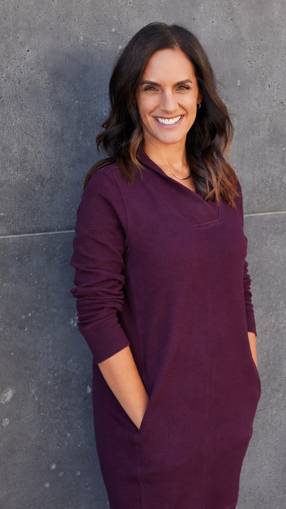A woman with dark brown hair in loose waves, smiling, wearing a purple long-sleeve sweater and standing against a gray concrete wall.