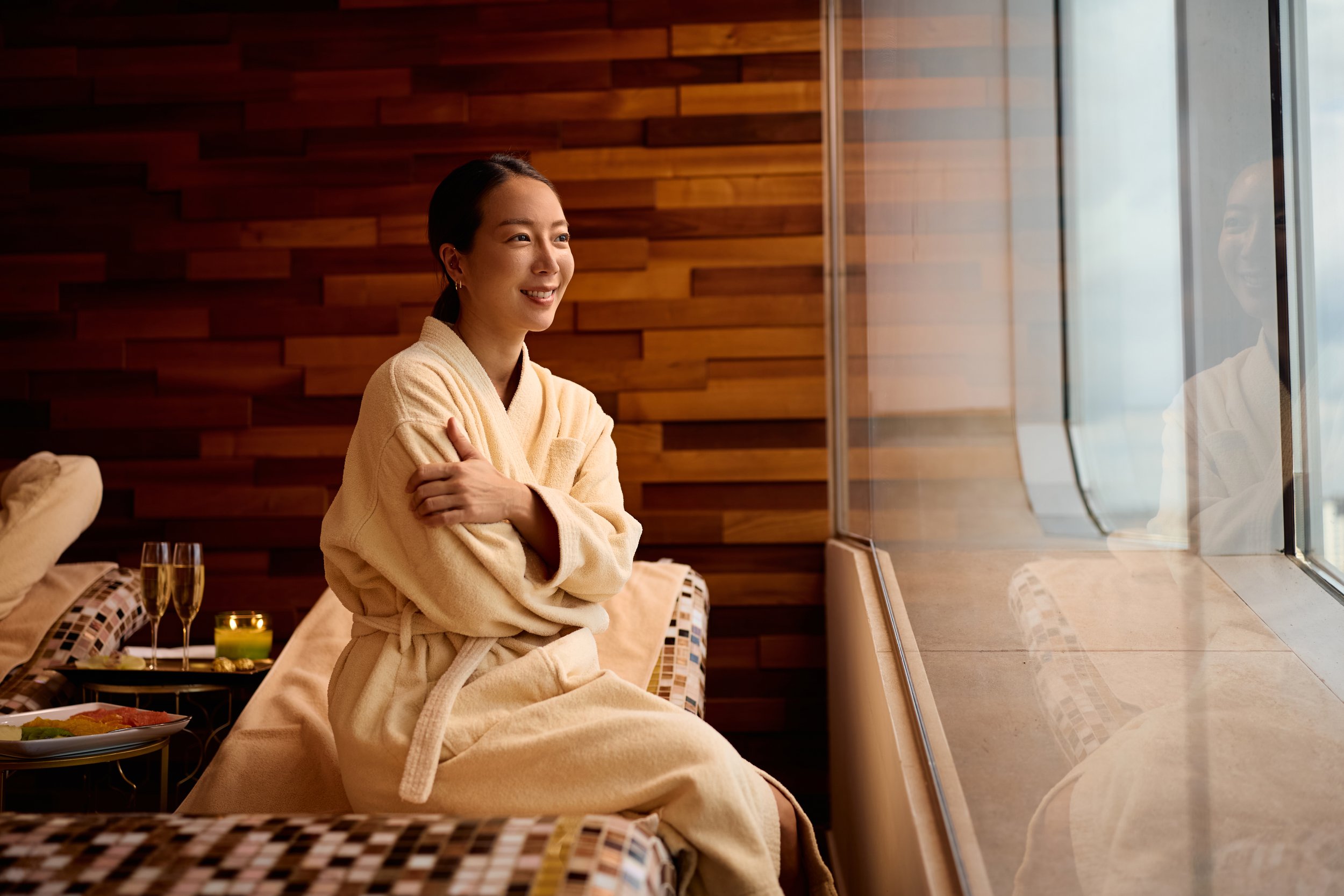 A young woman wearing a beige bathrobe sitting by a large window in a cozy, wood-paneled room. She is smiling, with arms crossed, looking outside. On the table nearby are two glasses of champagne, a lit candle, and a platter of food, suggesting a spa or relaxation setting.