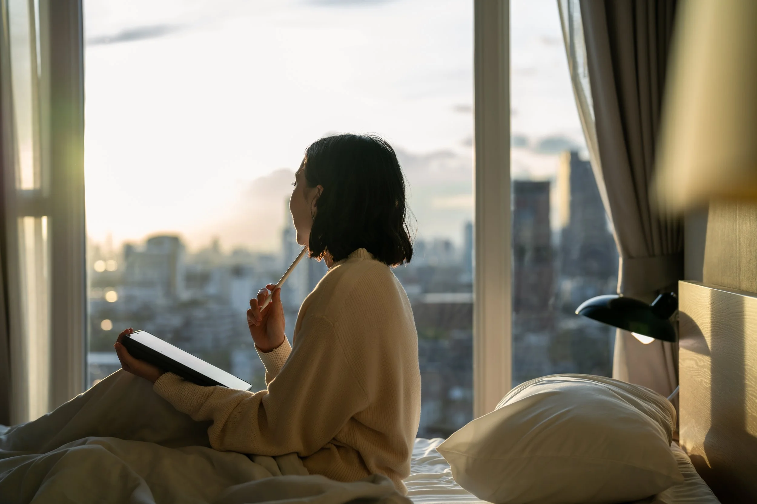 A woman with shoulder-length dark hair, wearing a cream-colored sweater, sitting on a bed near a large window during sunrise or sunset. She is holding a tablet in one hand and a stylus or pen near her lips. The city skyline is visible outside the window, with soft natural light illuminating the scene.