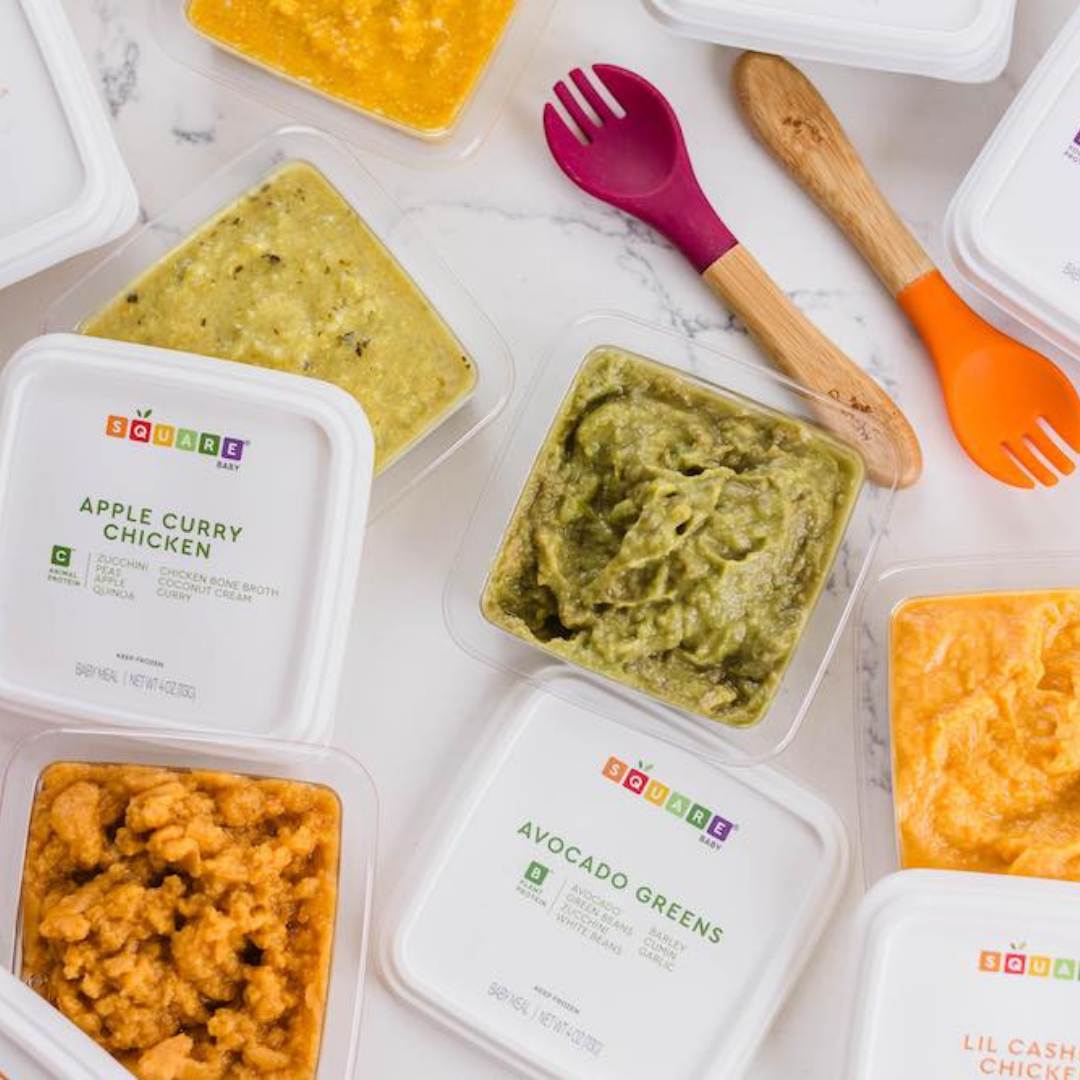 Containers of baby food with flavors including apple curry chicken, avocado greens, and other purees, with colorful baby spoons and a white marble surface.